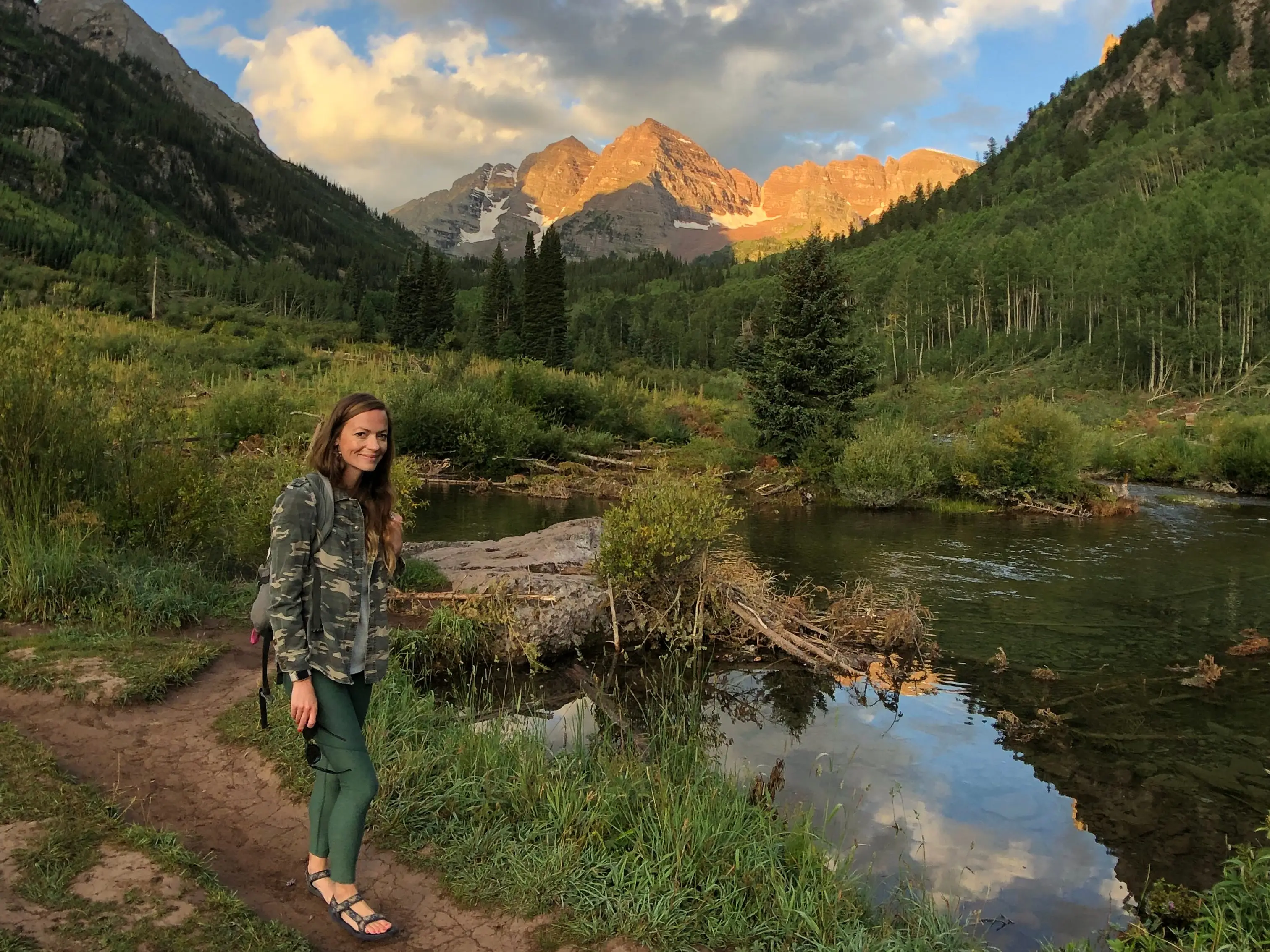 Emily stands on a path near a lake and trees with mountains in the distance.
