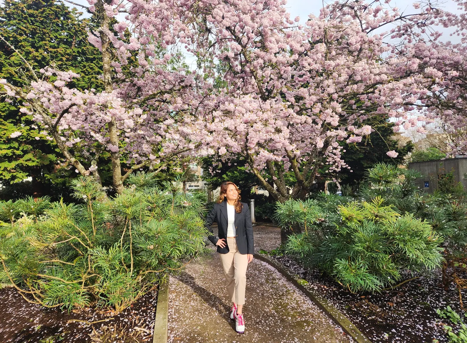 The writer in a blazer and pants, walking through a garden with trees and pink flowers.