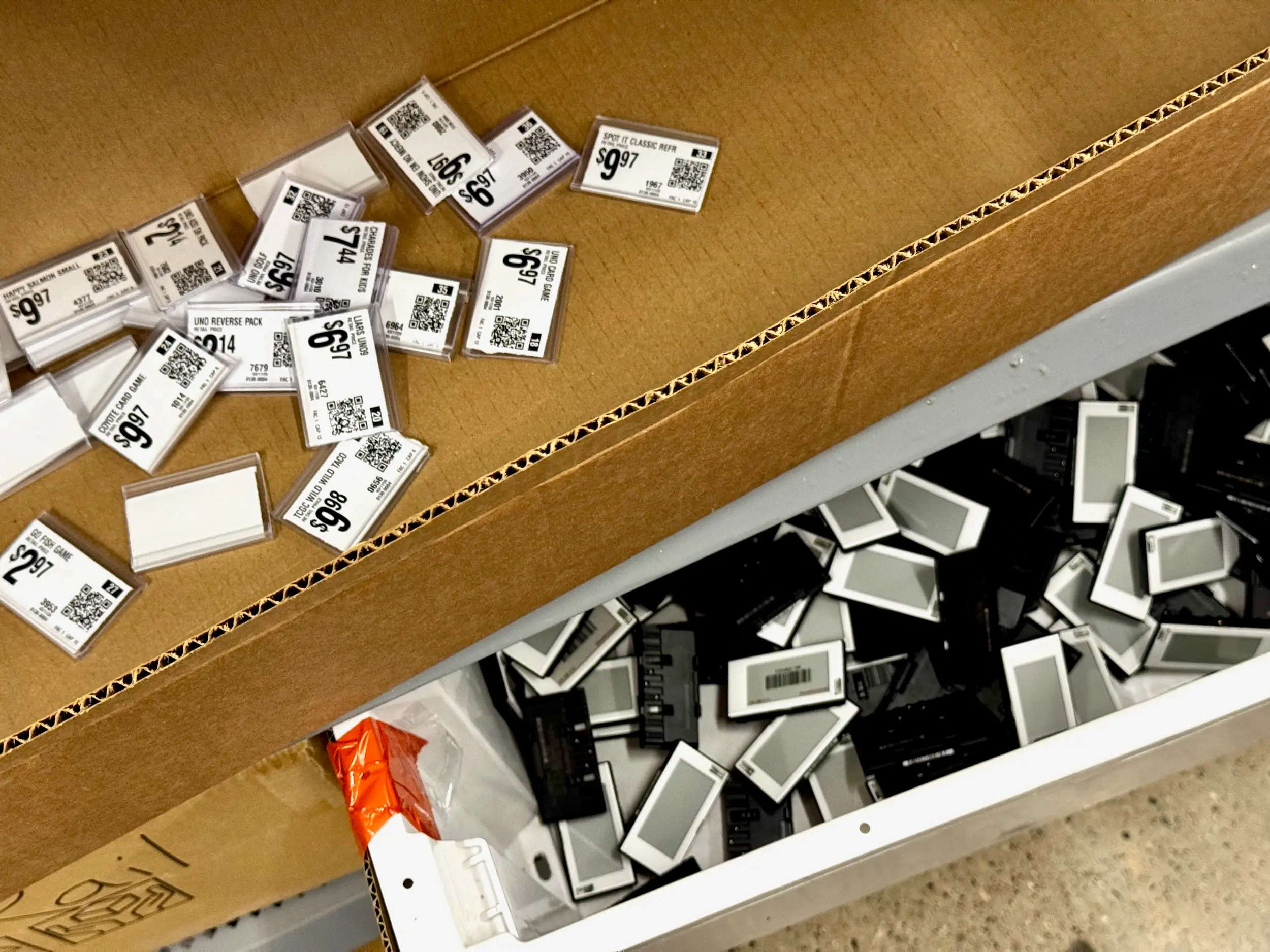 A cardboard box contains scattered electronic shelf labels alongside a bin filled with paper tags.