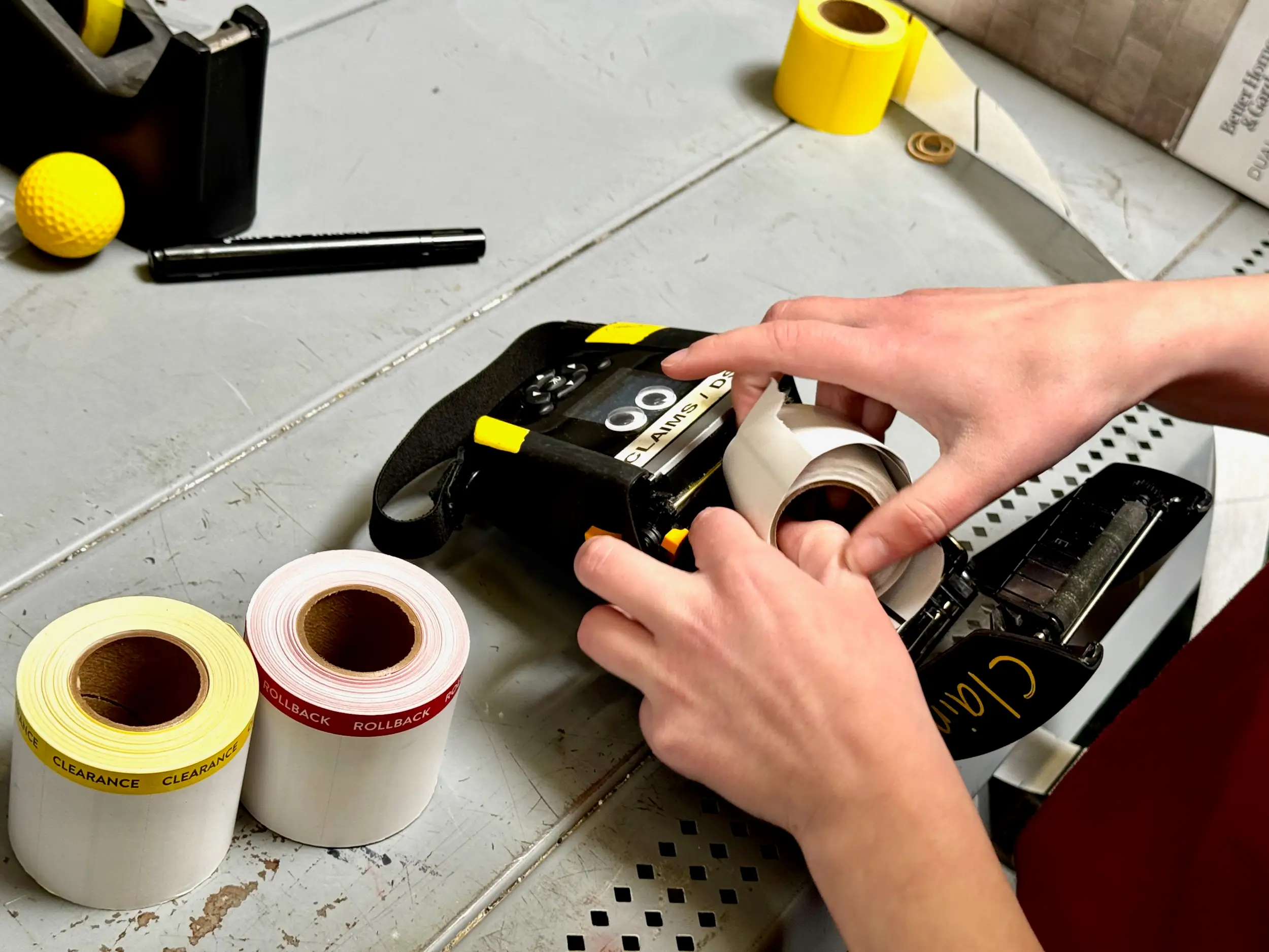 Hands load a roll of labels into a handheld printer beside rolls marked clearance and rollback.