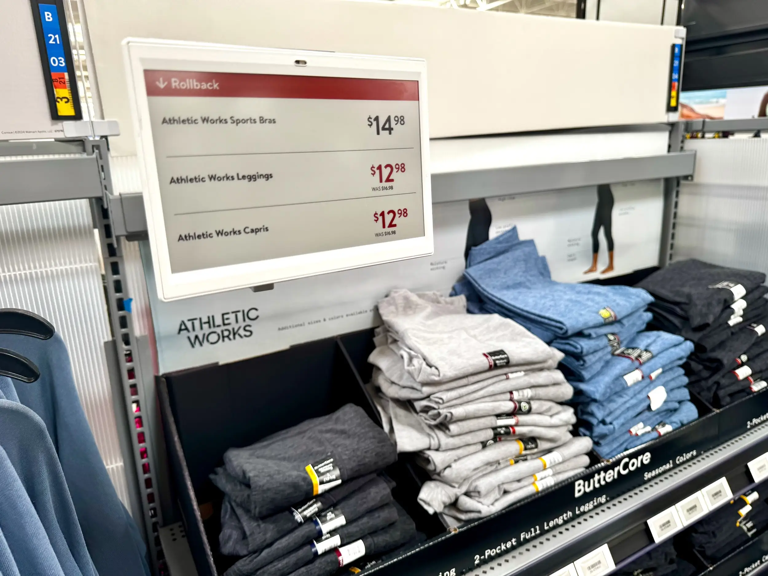 Folded athletic clothing sits beneath a digital sign showing rollback prices for bras, leggings, and capris.