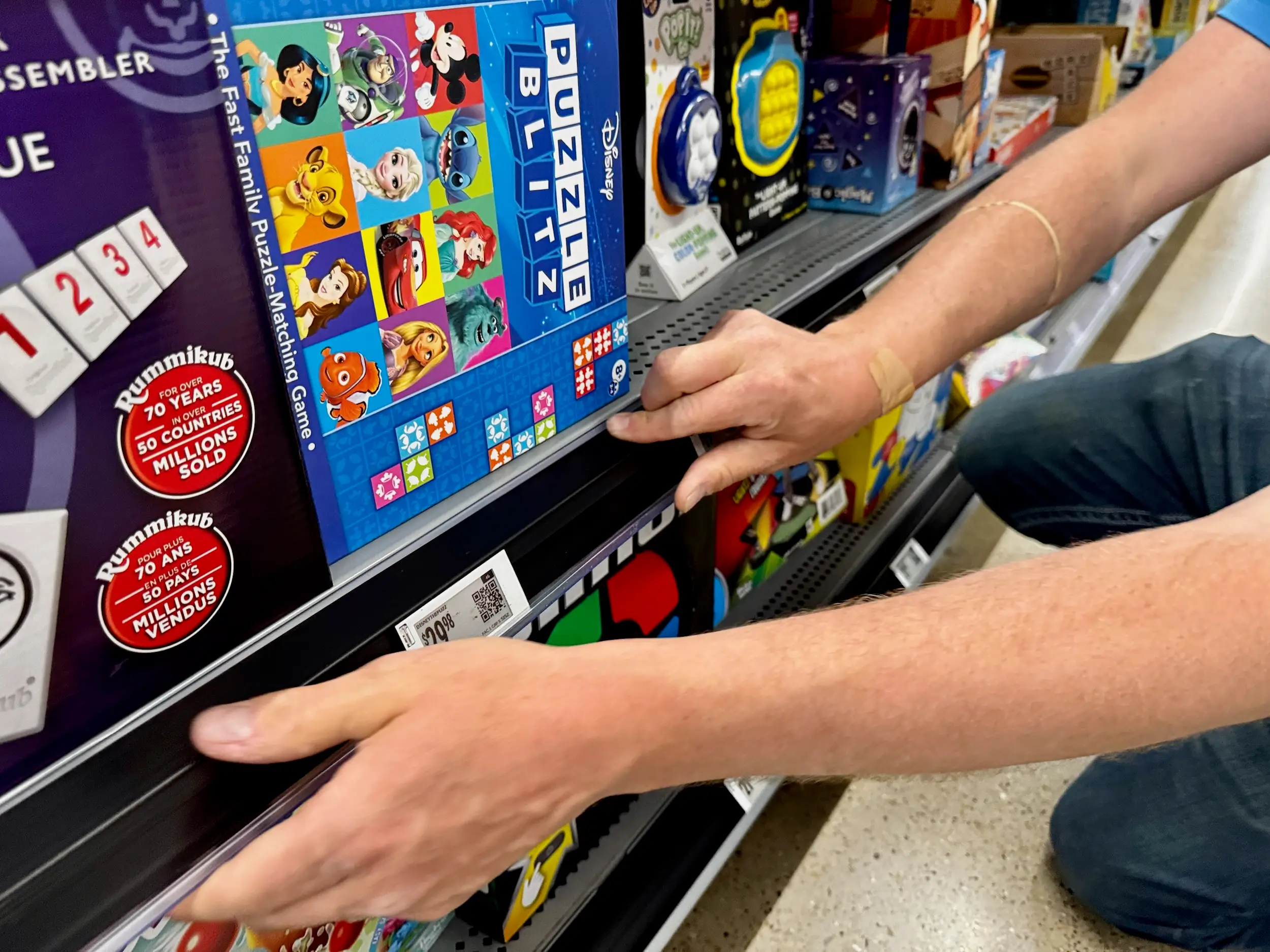 A worker adjusts electronic shelf labels beneath board games like Disney puzzles and Rummikub.