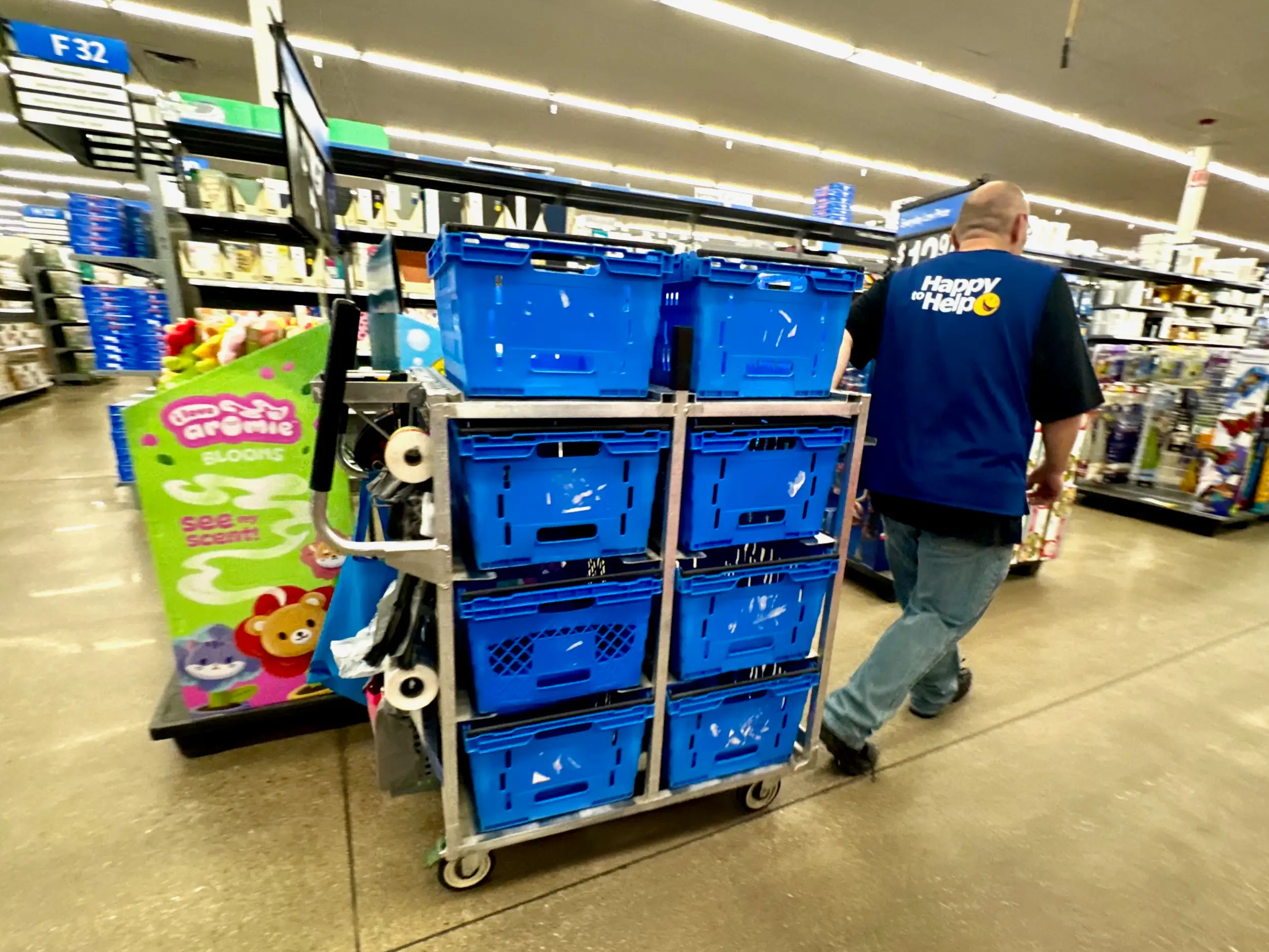 A worker pushes a cart stacked with blue bins down a store aisle filled with merchandise.
