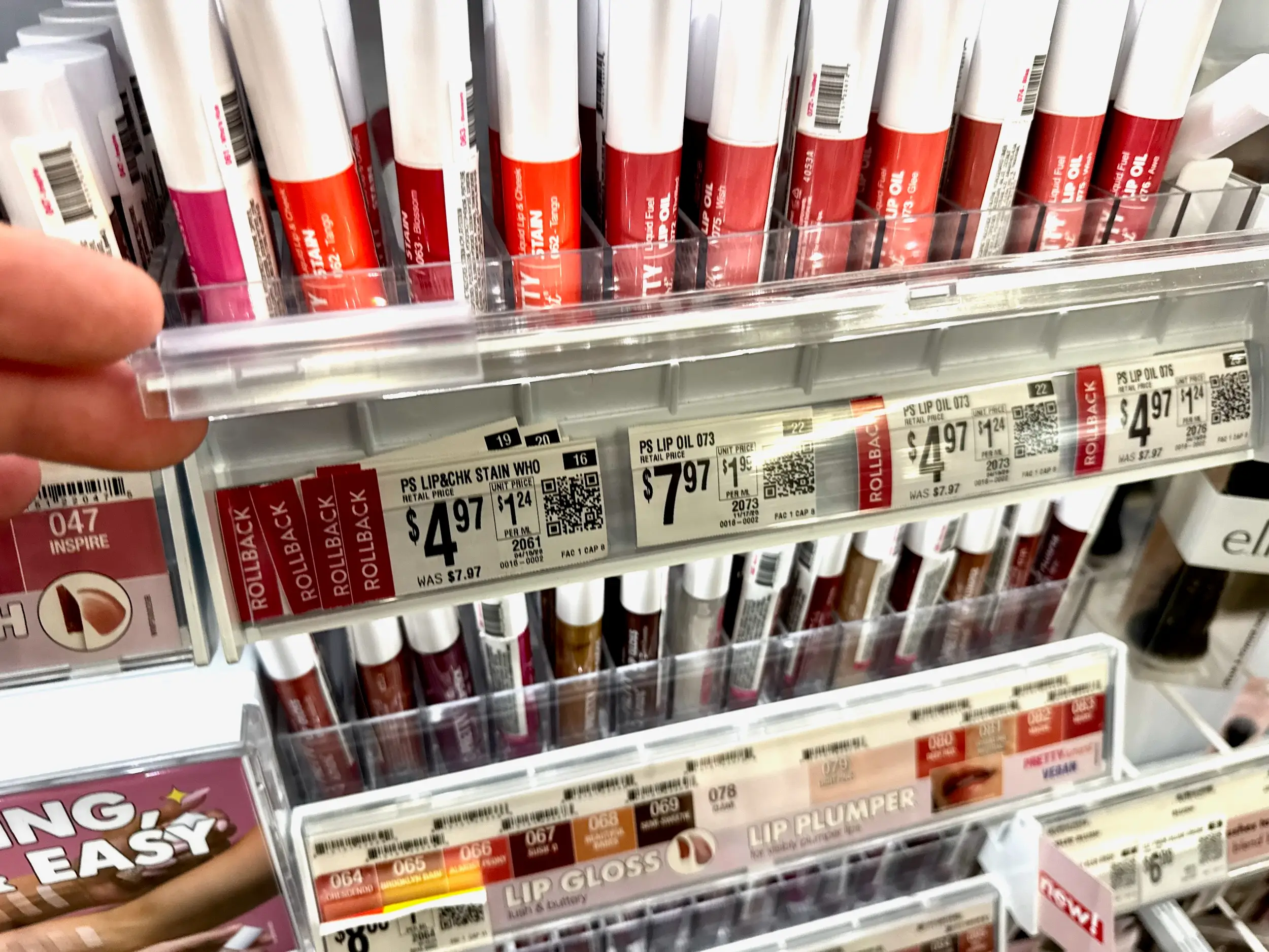 A close-up shows lip products on a shelf with rollback price tags and a hand adjusting the display.