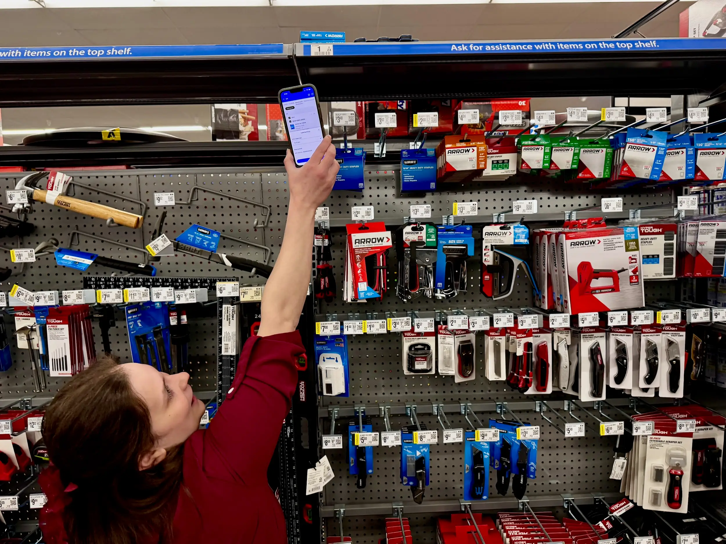 A store worker reaches up with a smartphone to scan a shelf code in a hardware aisle filled with tools.