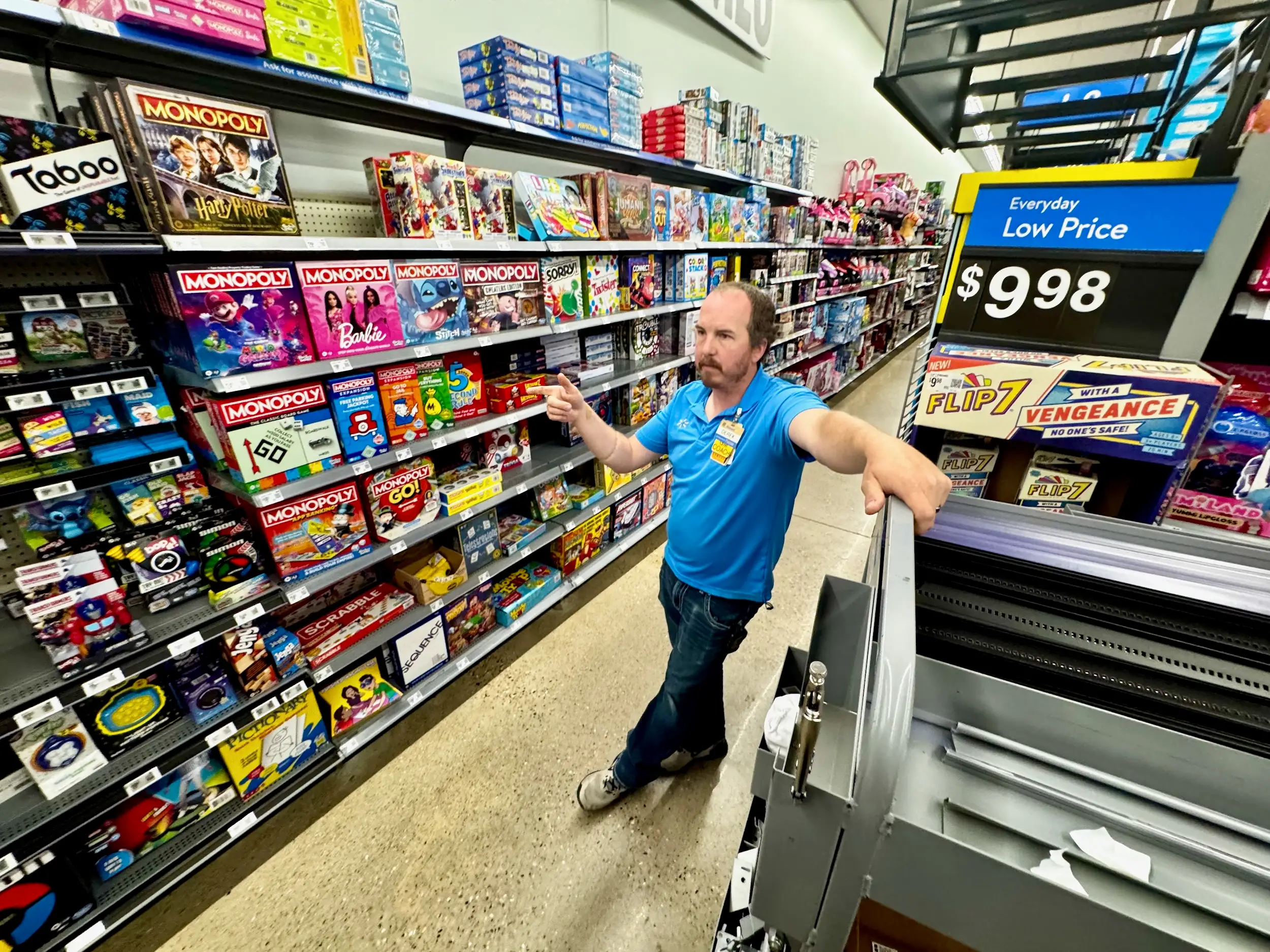 A store employee points toward board games on a shelf, including Monopoly and Scrabble.