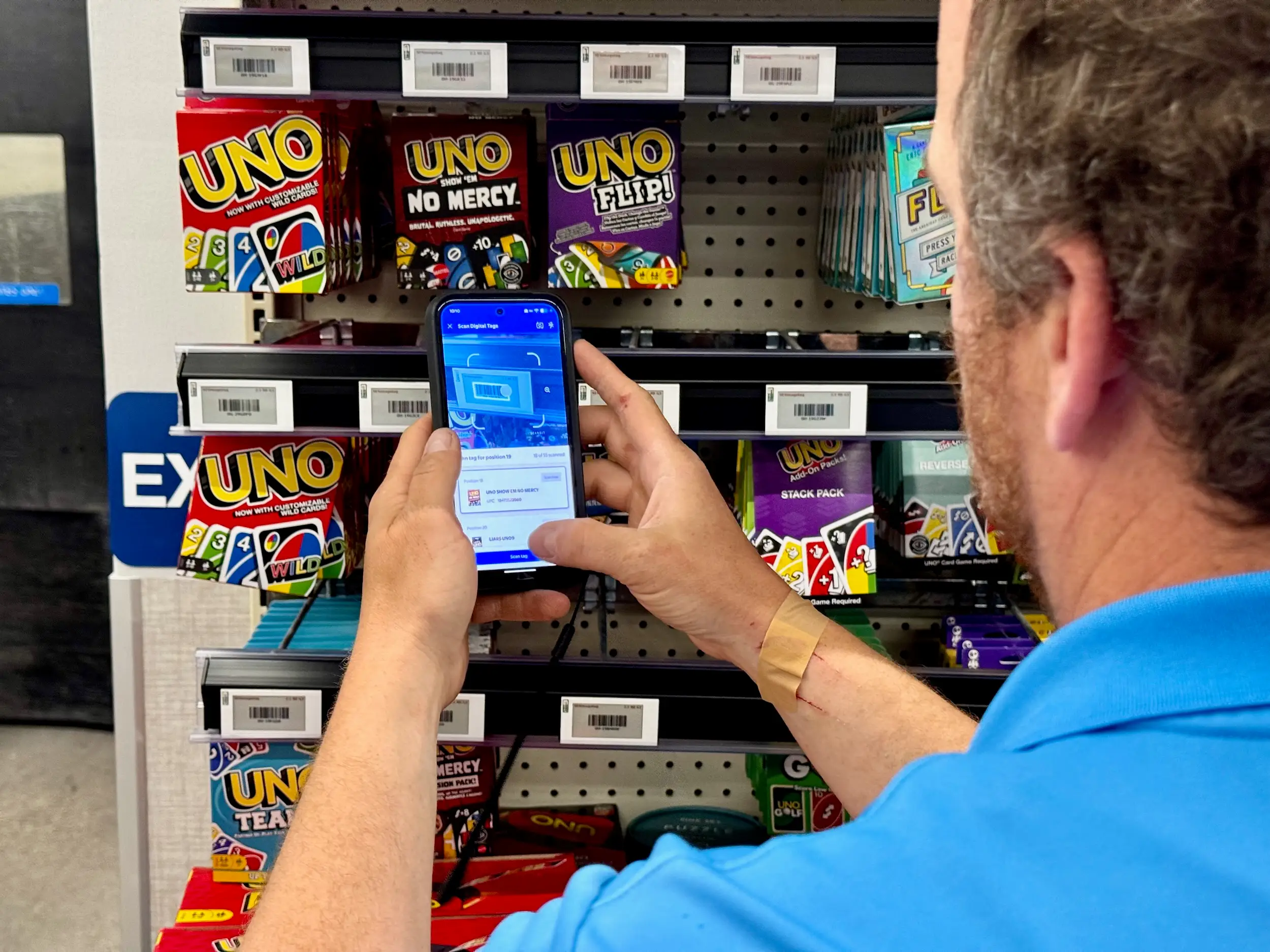 A close-up shows a person scanning a shelf label with a smartphone in front of Uno card game boxes.