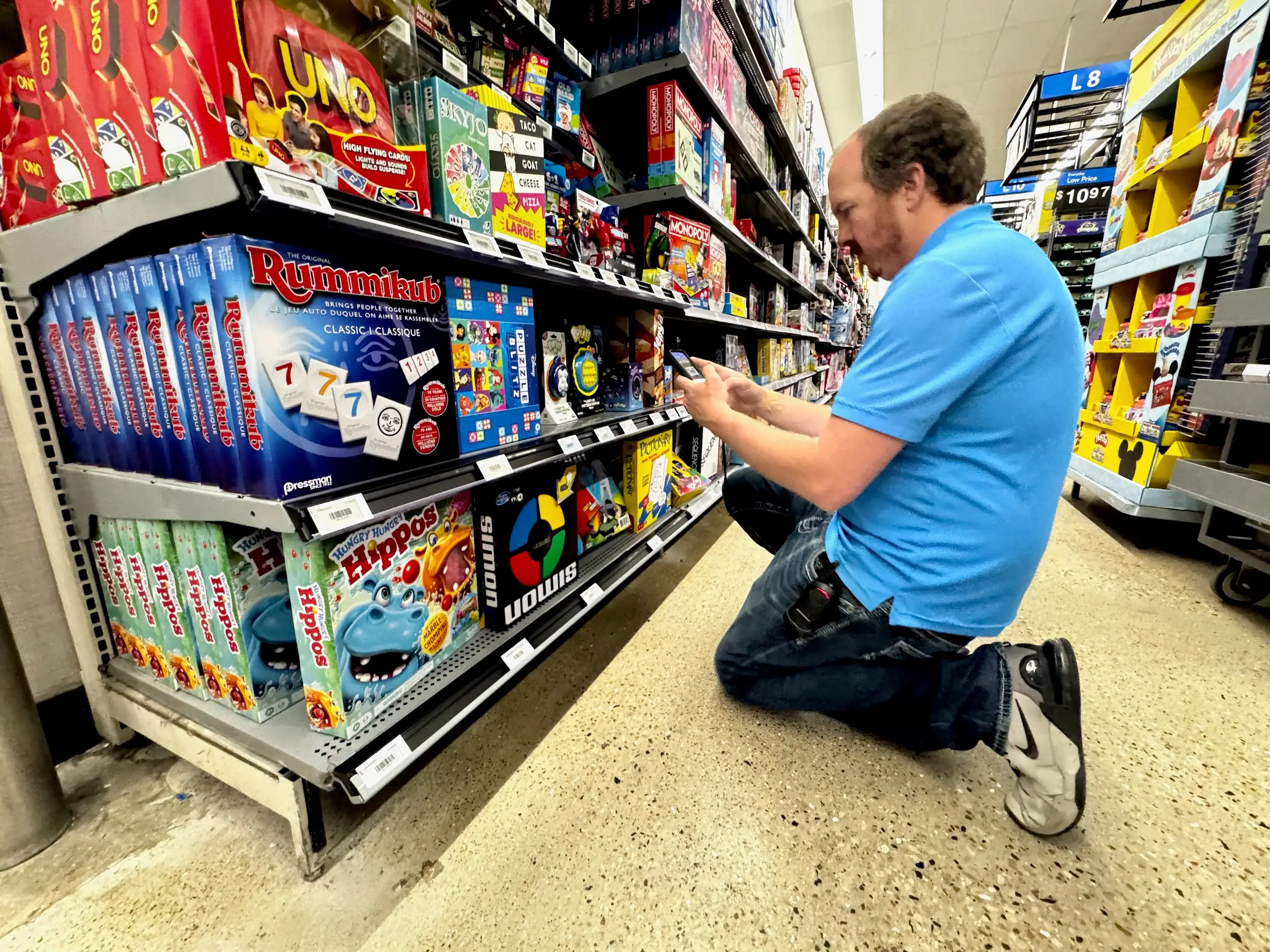 A worker kneels in a toy aisle using a smartphone to scan or manage items on a shelf.