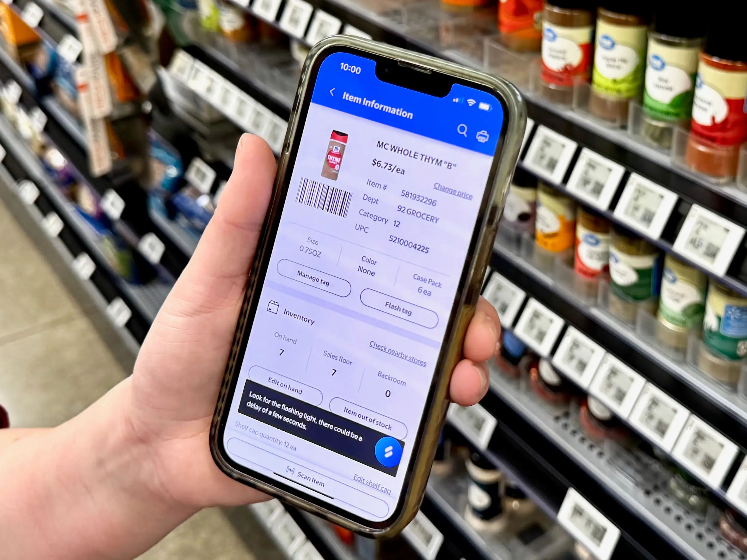 A hand holds a smartphone displaying item information for thyme in front of a spice aisle with digital price tags.