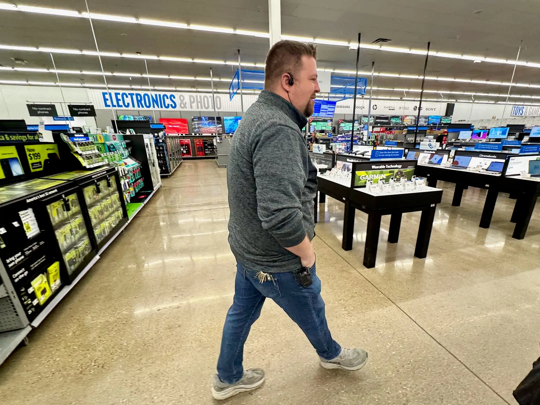 A store employee walks through an electronics section with tables of devices and overhead department signs.