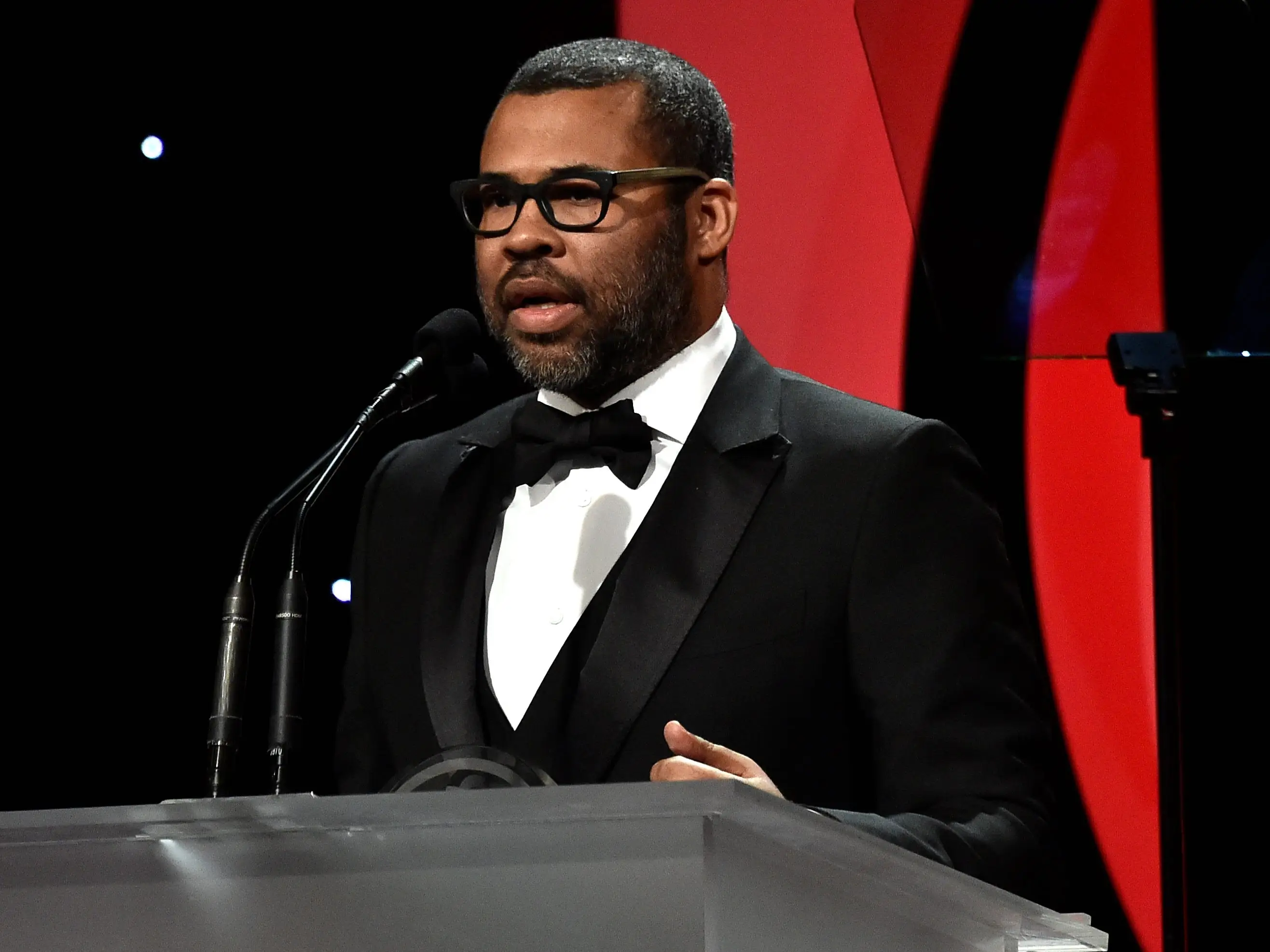 Jordan Peele in a suit at a podium speaking into a microphone