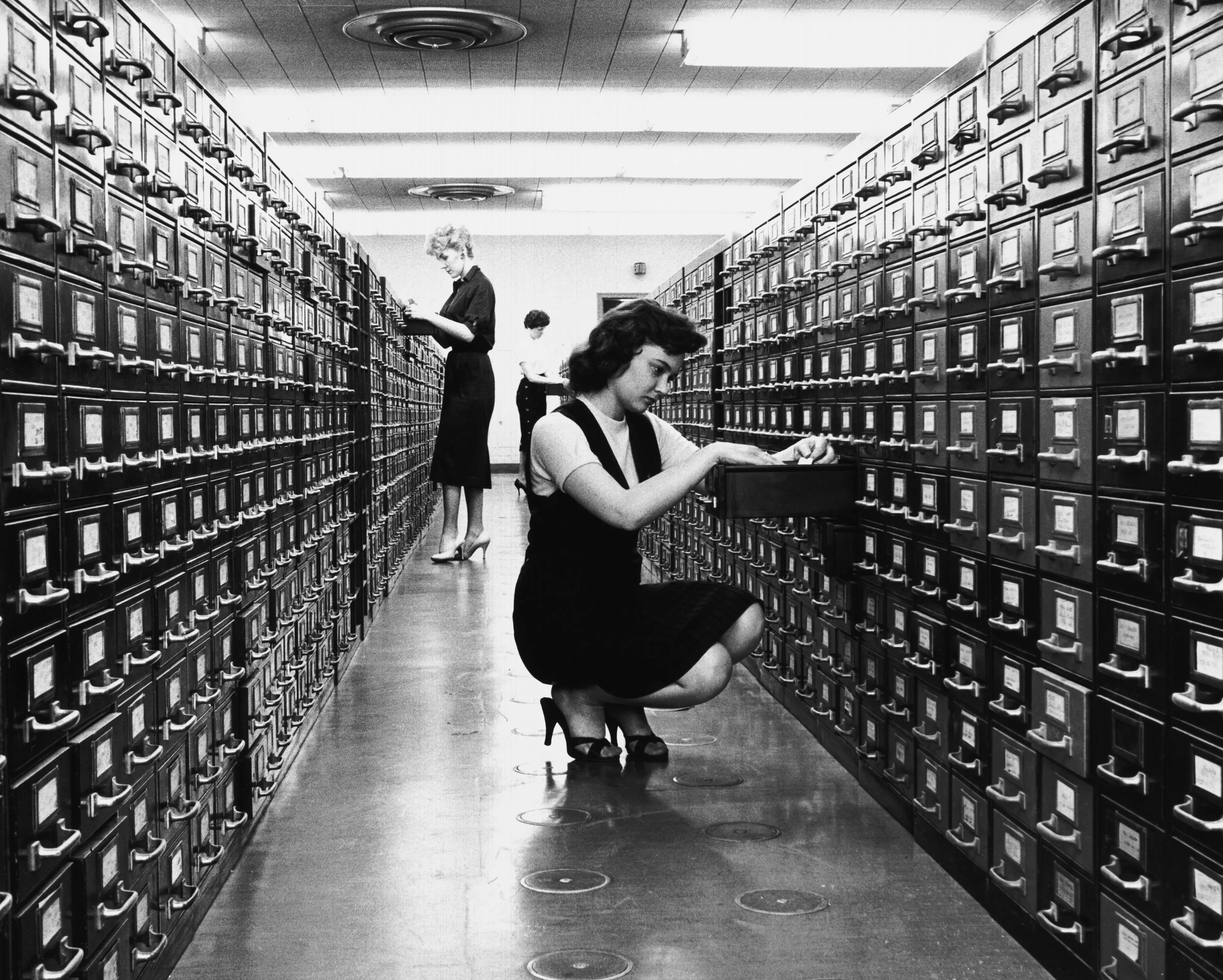 Three women, standing between two rows of filing cabinets, are consulting file cards and collecting information for the preparation of the new 'All Canadian Atlas', Ottawa, Ontario, Canada, 1959.