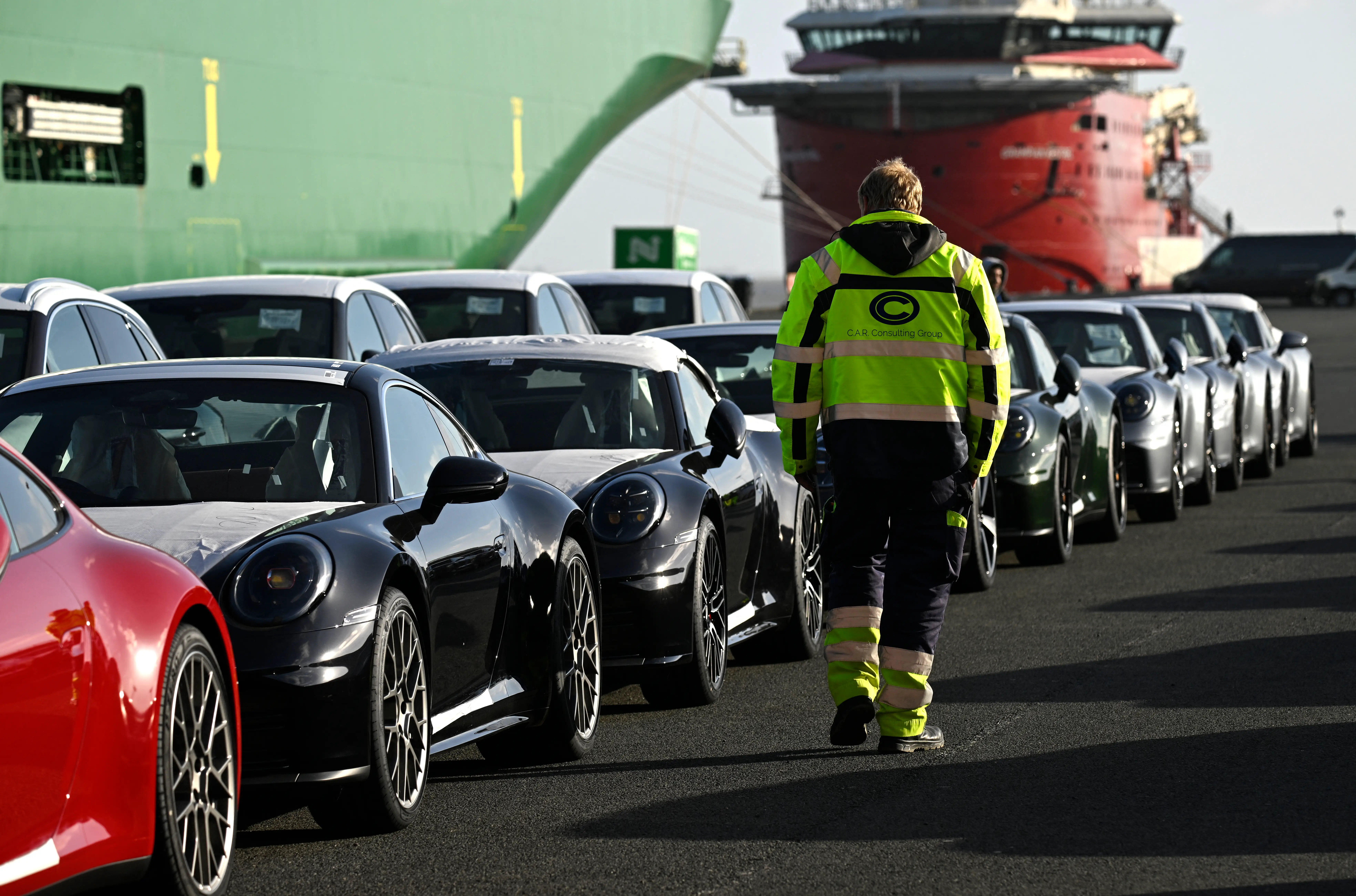 A man in an electric green jacket walks past a lineup of Porsche vehicles at a shipping port.