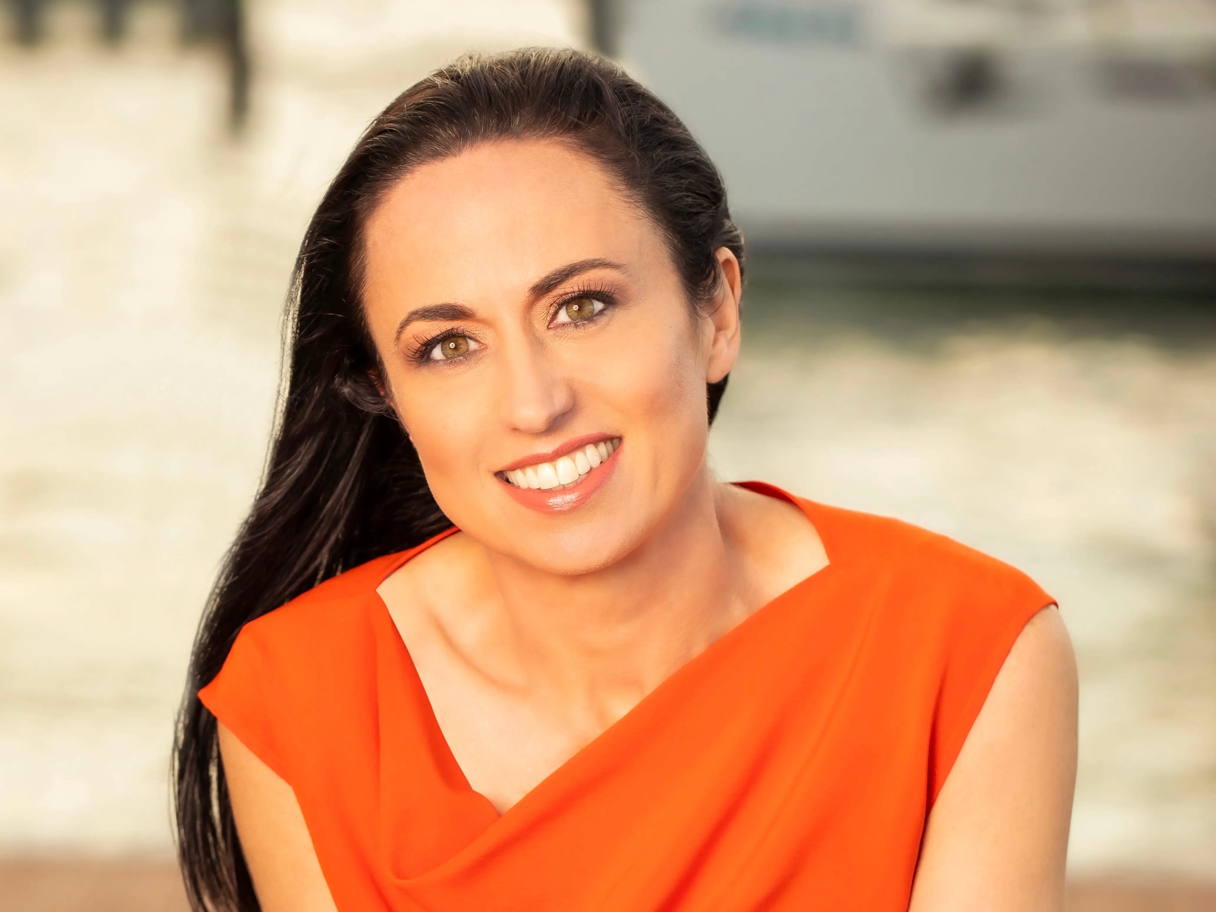 Leaning forward with hands clasped, a woman in an orange top smiles at the camera against a softly blurred outdoor background.