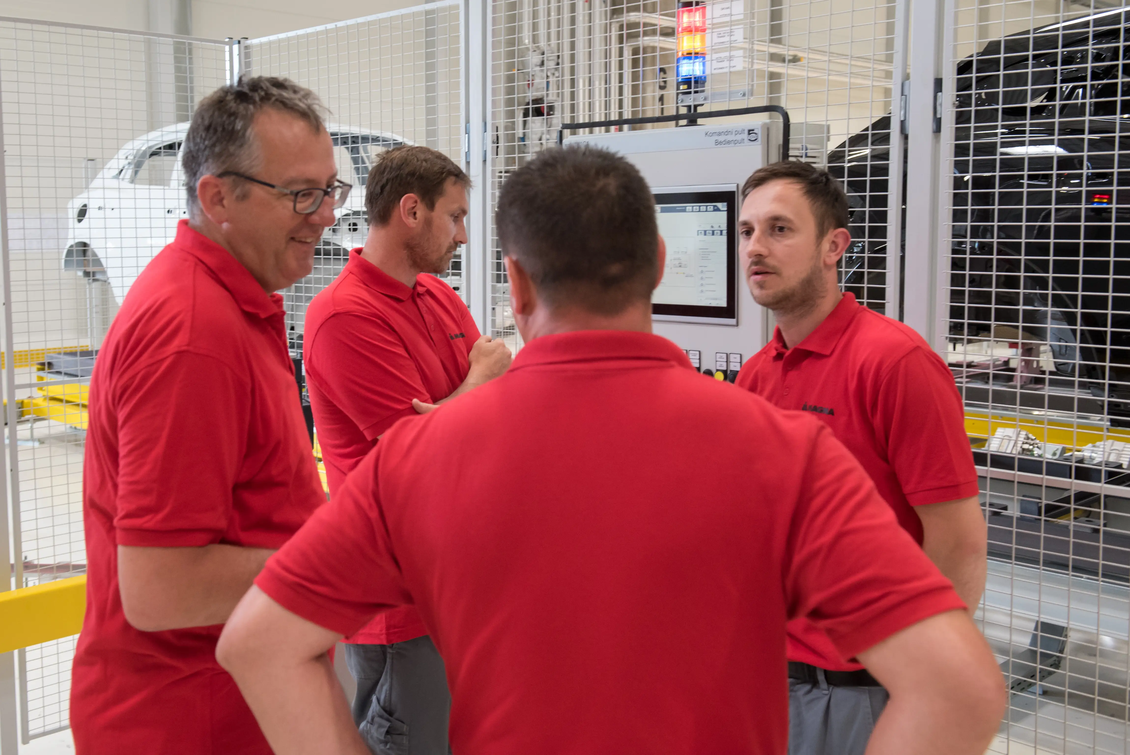 Four workers in red polo shirts stand together at a Magna factory next to a work station.