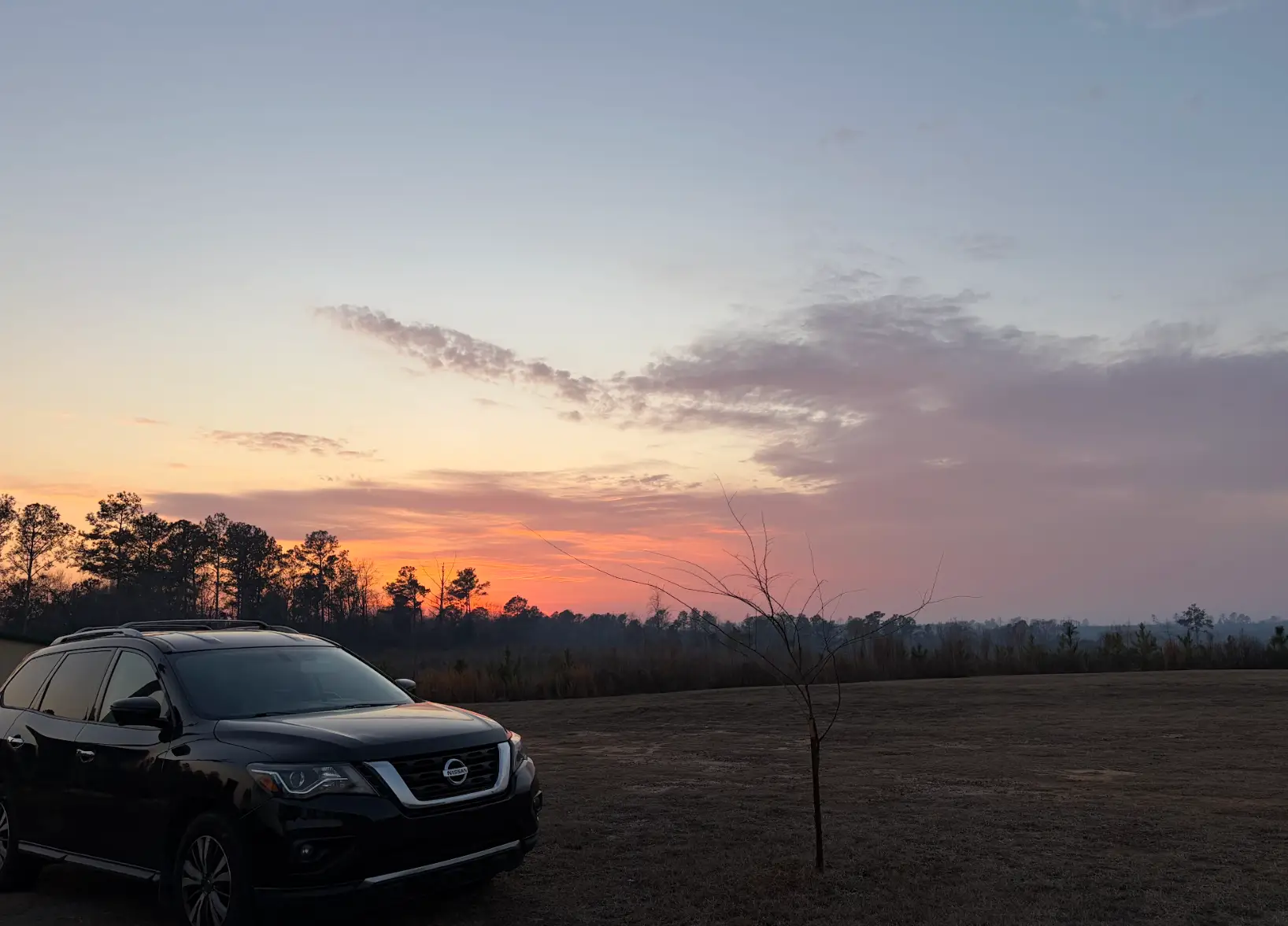 A car in the middle of a rural field, with the sun setting in the background.