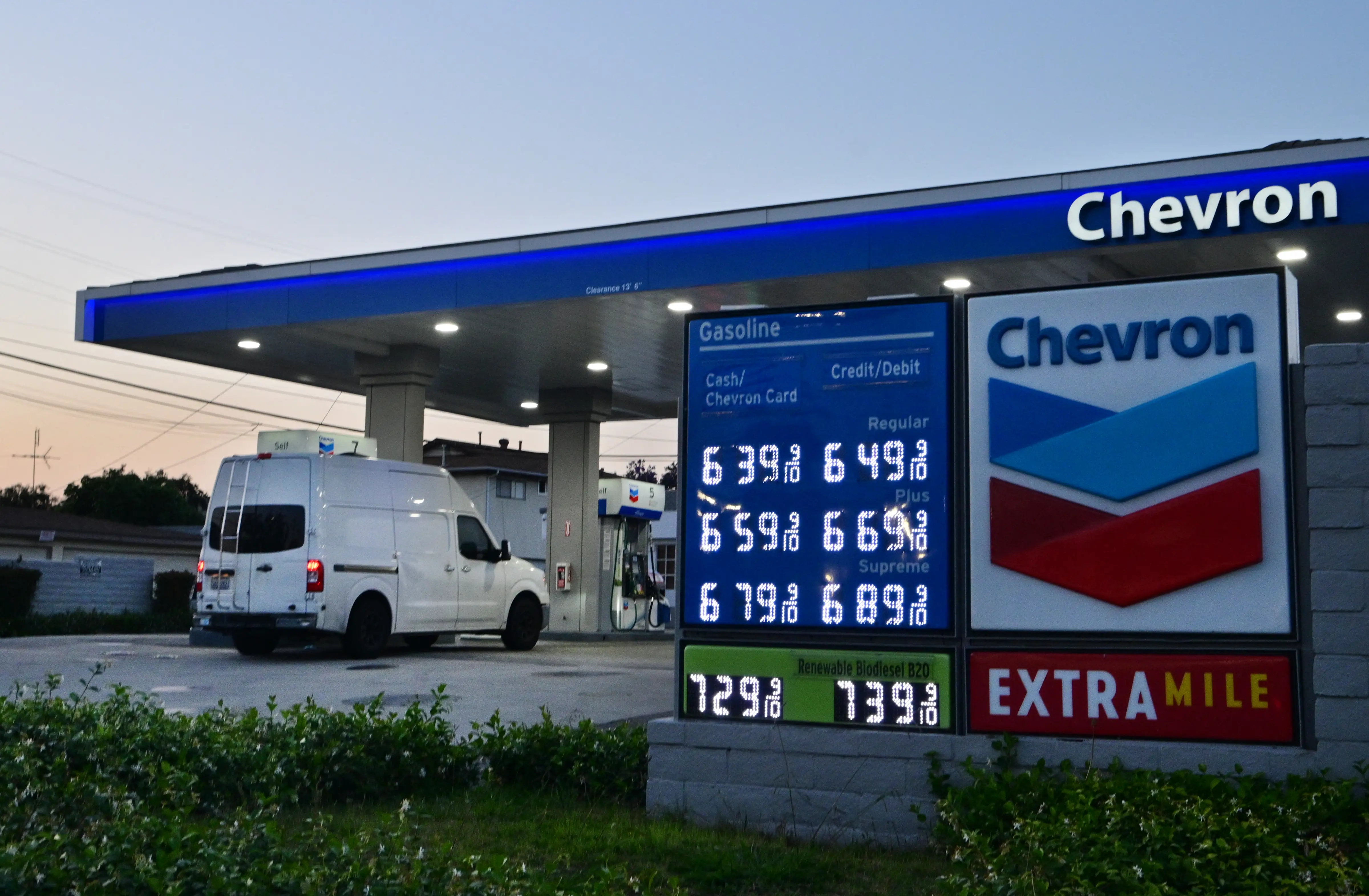 A panel van drives up to a Chevron gas station's pump at dusk. A lit-up sign advertises prices ranging from $6.39 to $7.39 a gallon.
