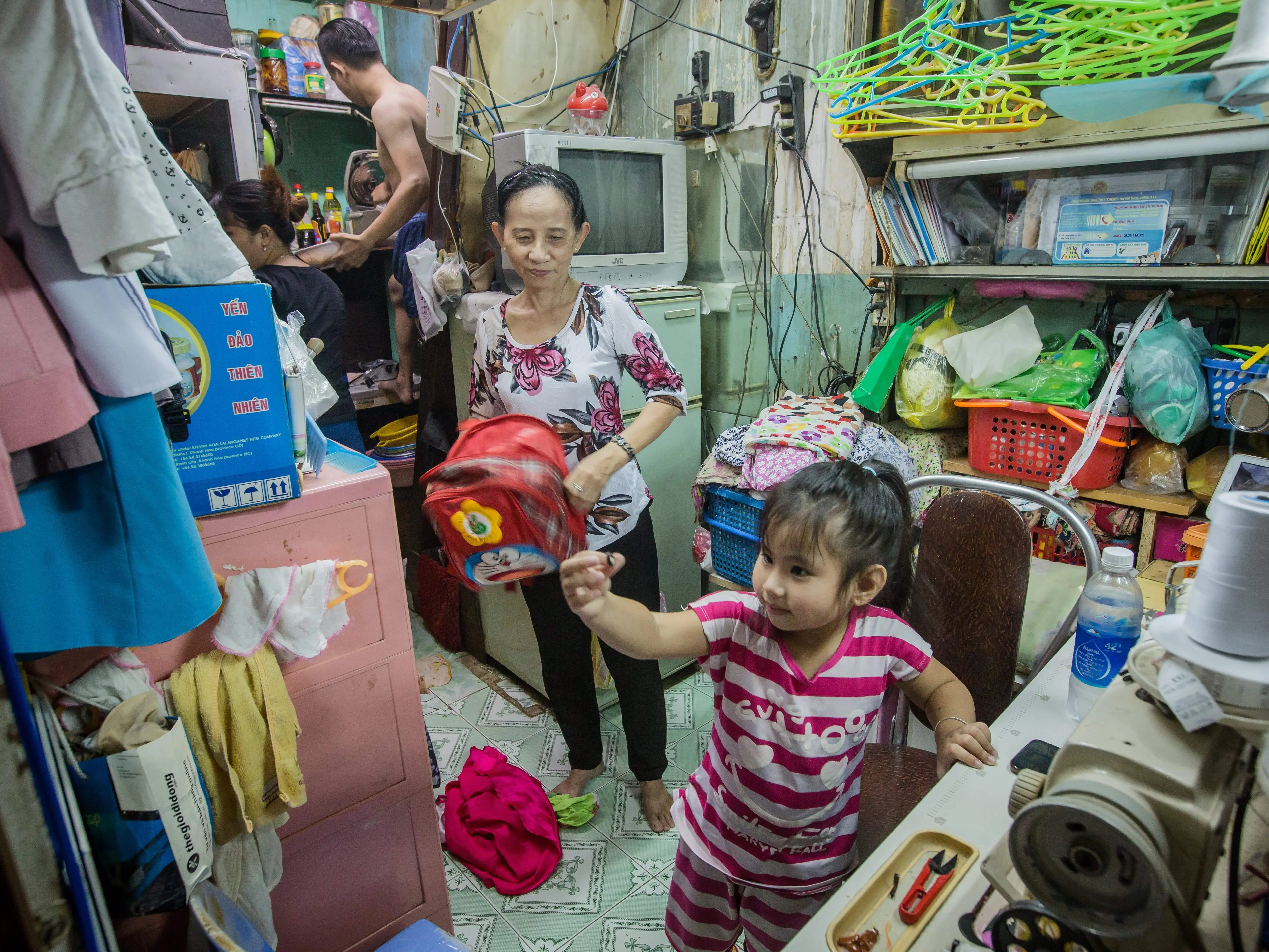 A Vietnamese woman holds a backpack and watches her daughter inside their cramped home.