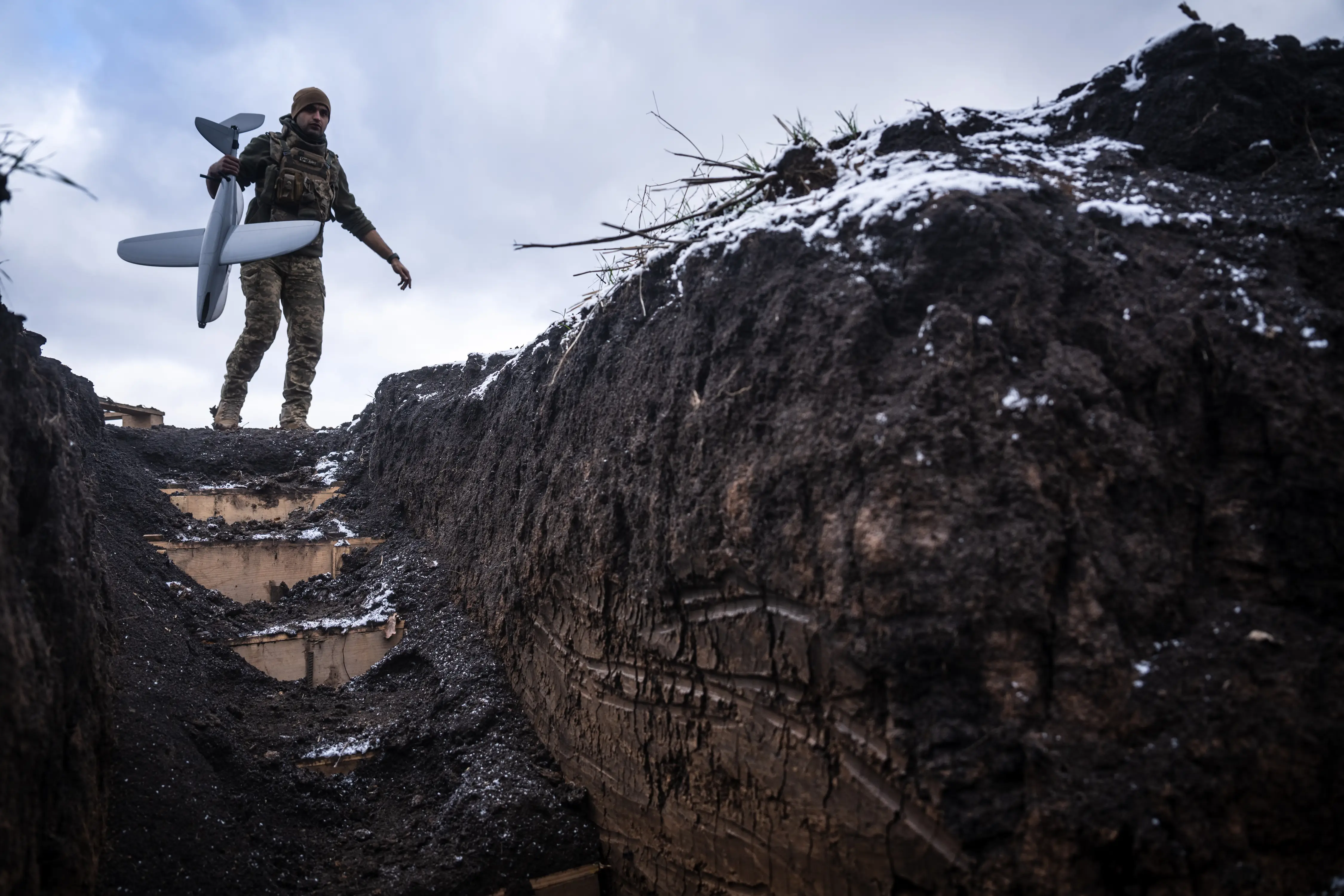 A man in camouflage walks down steps at the edge of a muddy trench, holding a grey drone