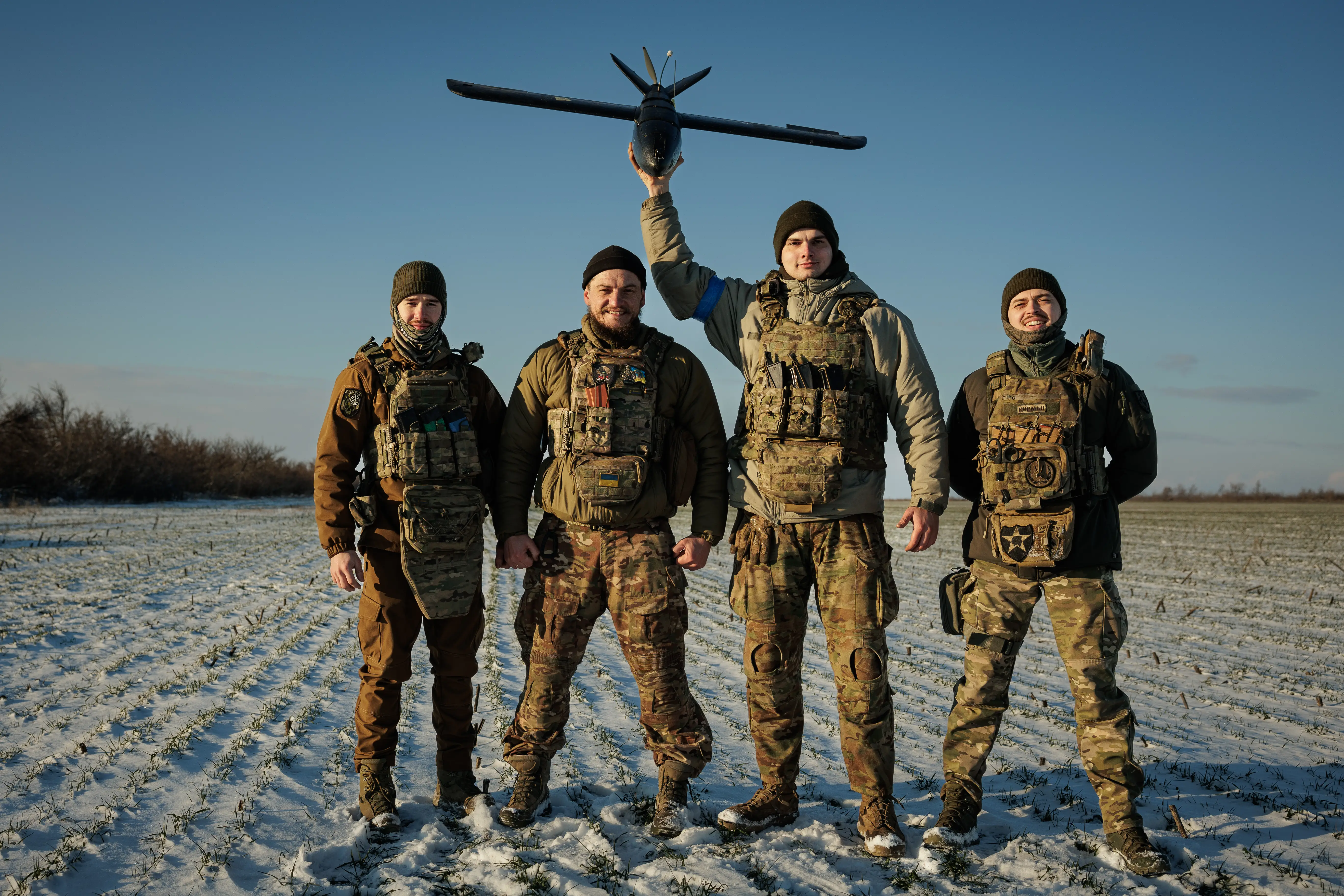 Four men in camouflage stand in a row in a sunny and snowy field with one holding a drone in the air