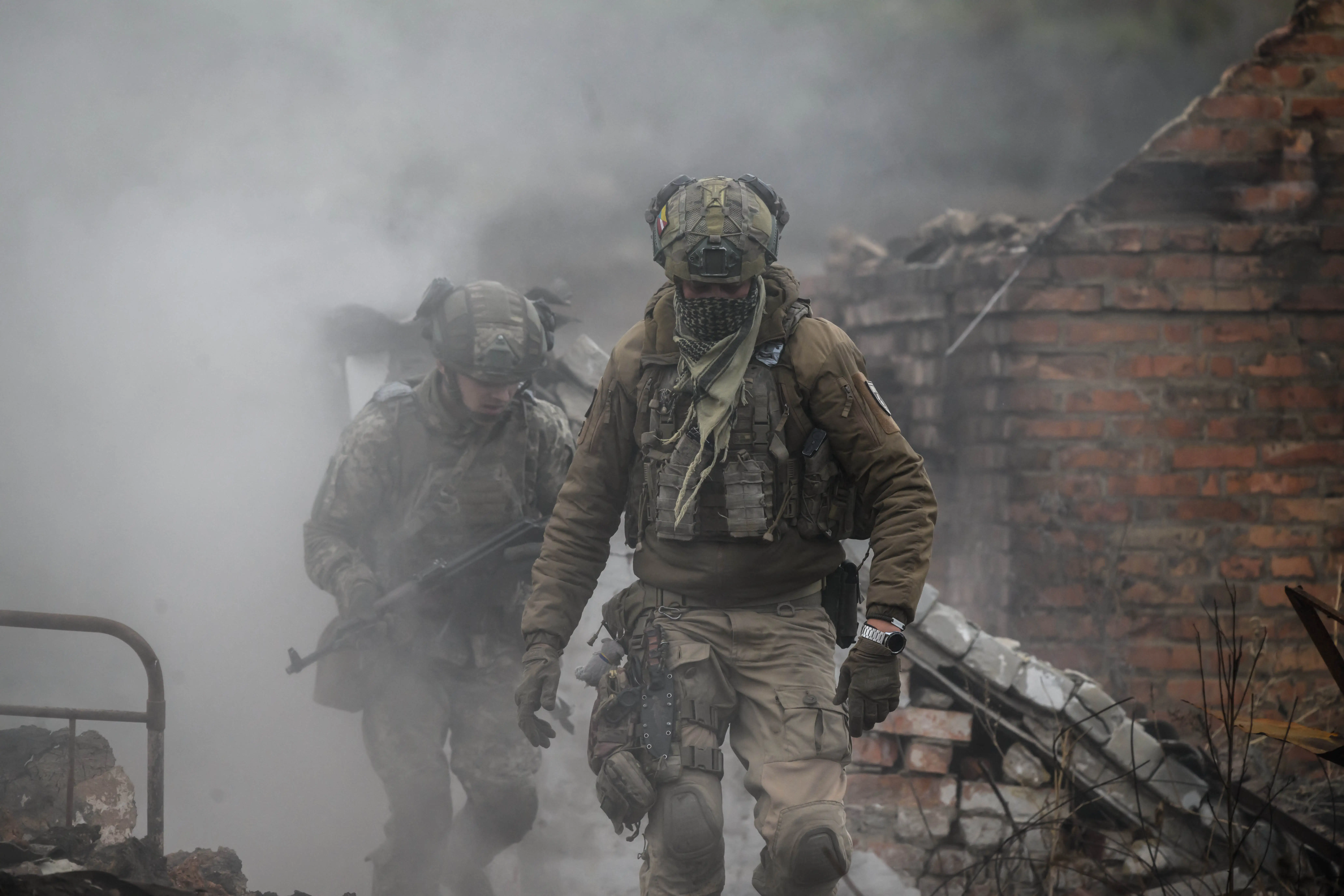 Two figures in khaki and helmets run with a red brick building and smoke in the background
