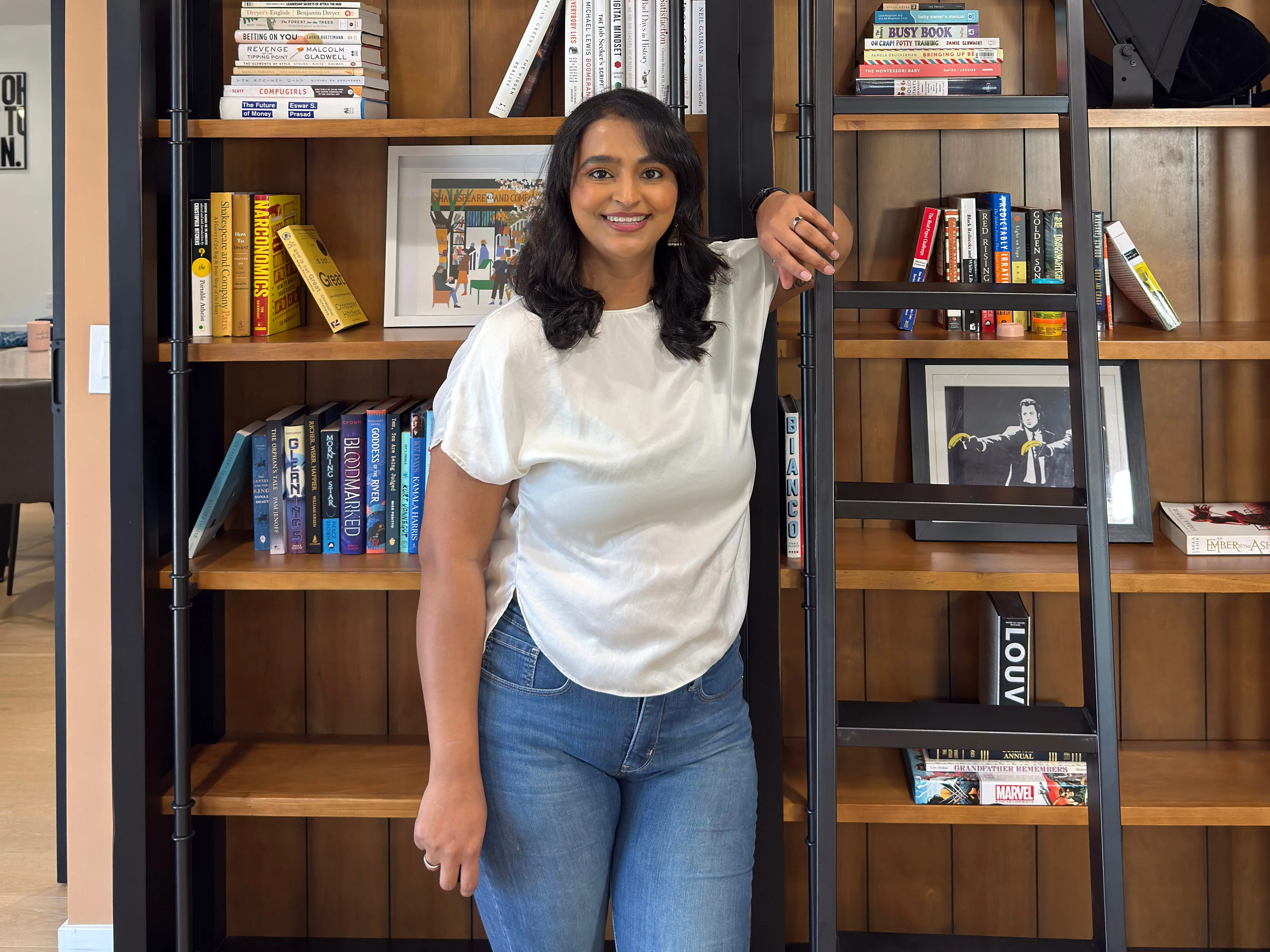 Tejal Rives is standing in front of a bookcase, leaning on a ladder.