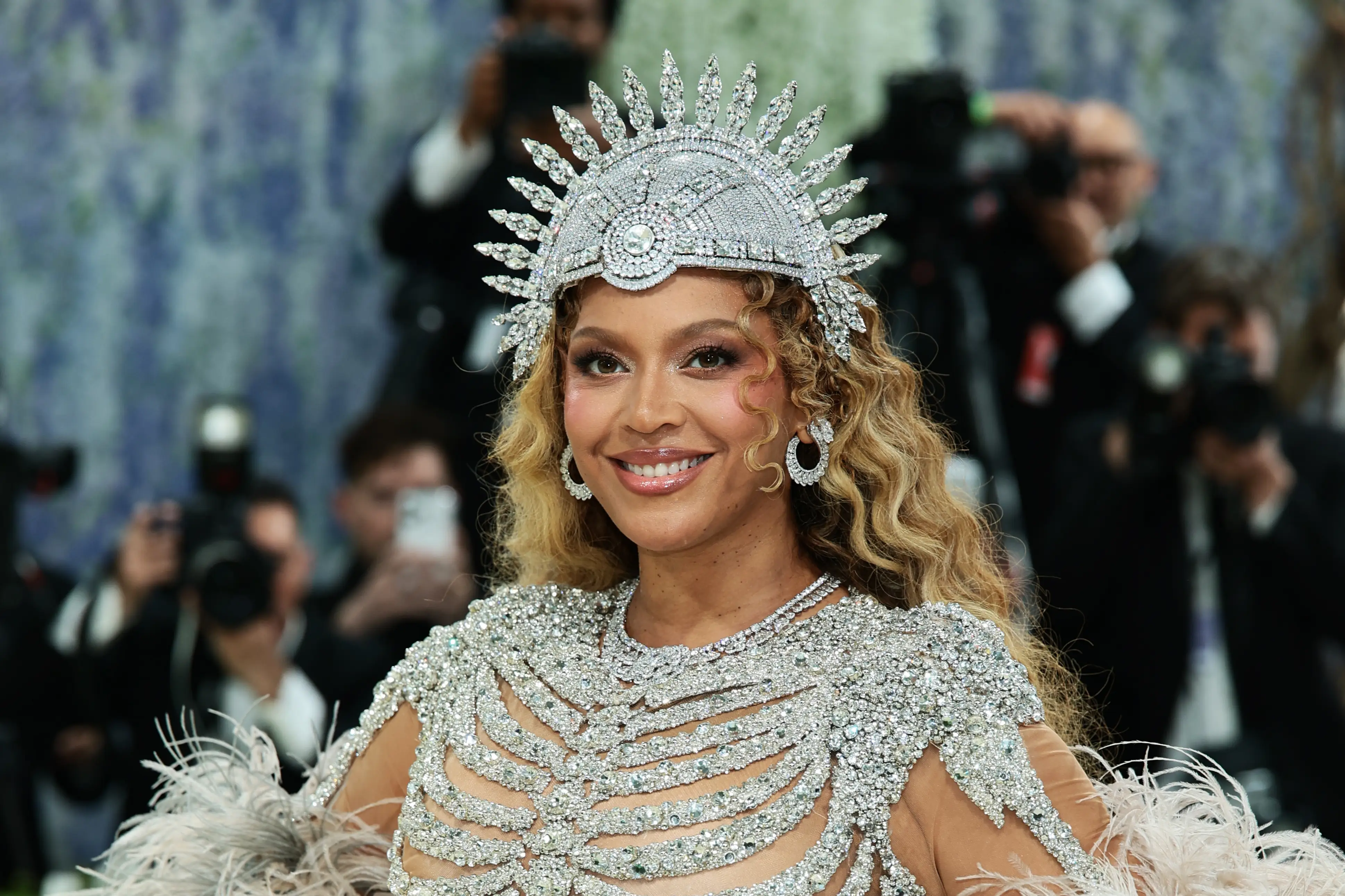 Beyoncé at the 2026 Met Gala, wearing a diamond-encrusted outfit and crown while paparazzi take photos.