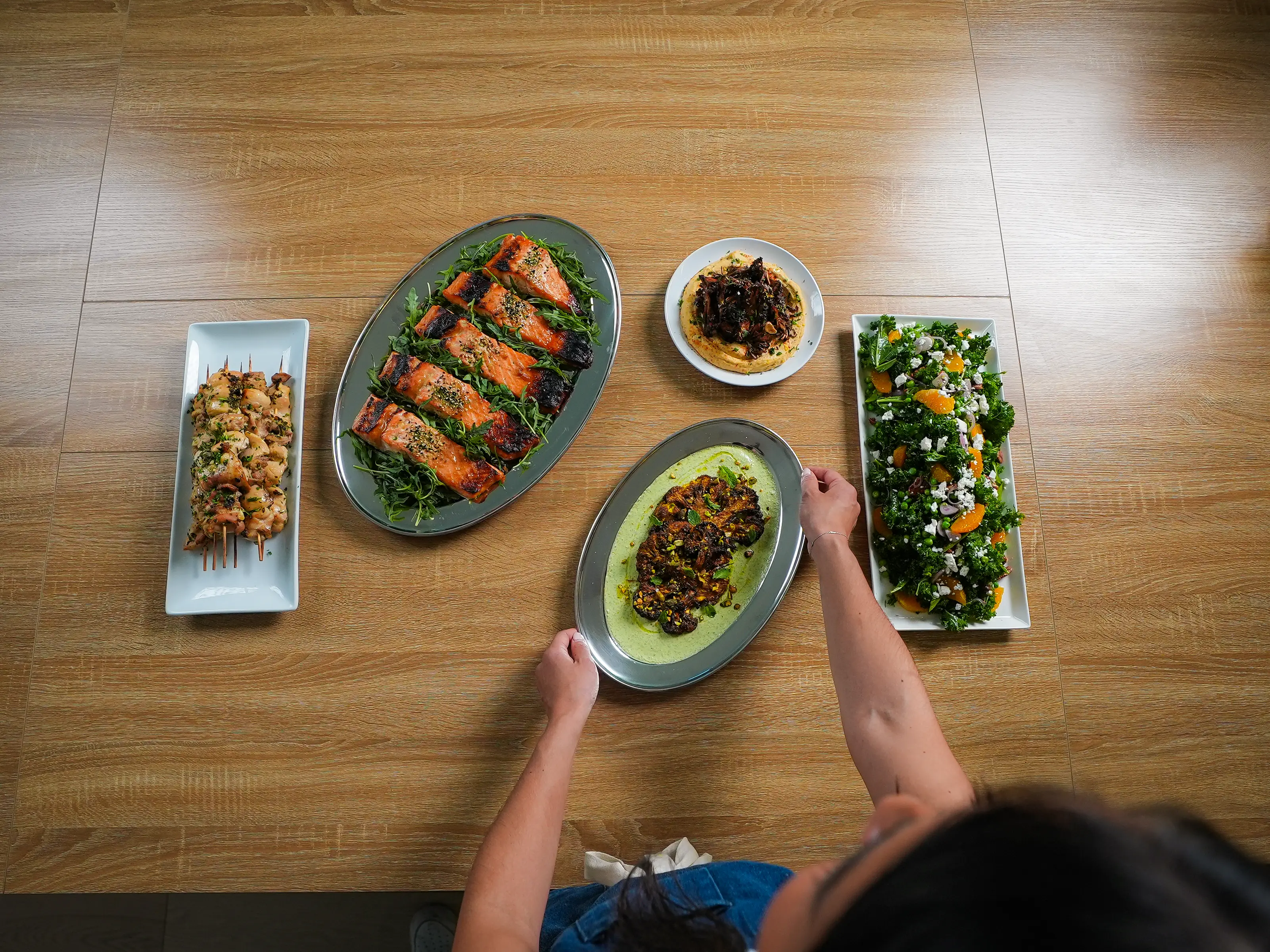 An overhead view shows a woman arranging several plated dishes, including grilled salmon, skewers, and salad, on a wooden table.