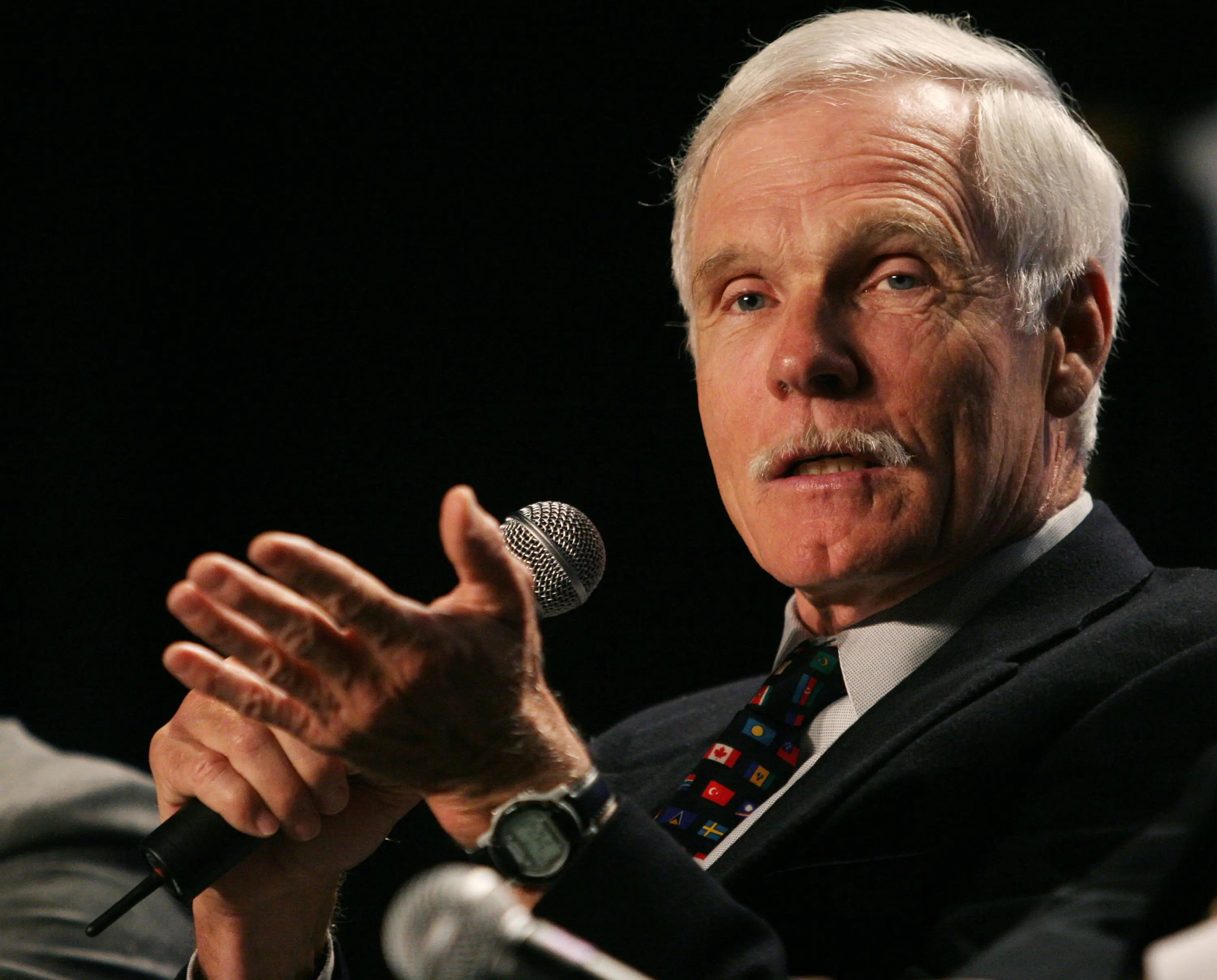 Ted Turner in a dark suit and tie talking into a microphone
