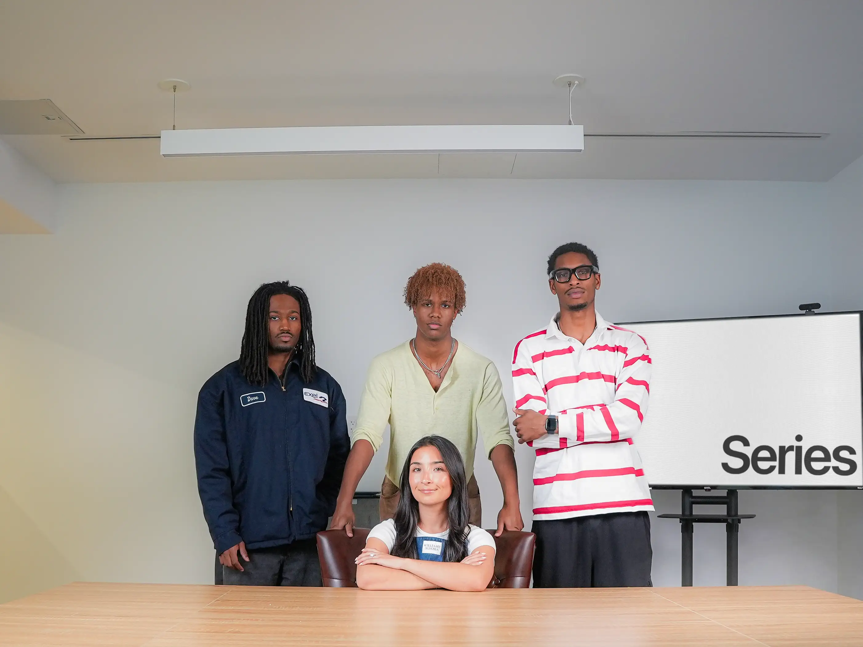 Four young adults pose around a conference table in a minimalist meeting room, with a screen displaying the word 
