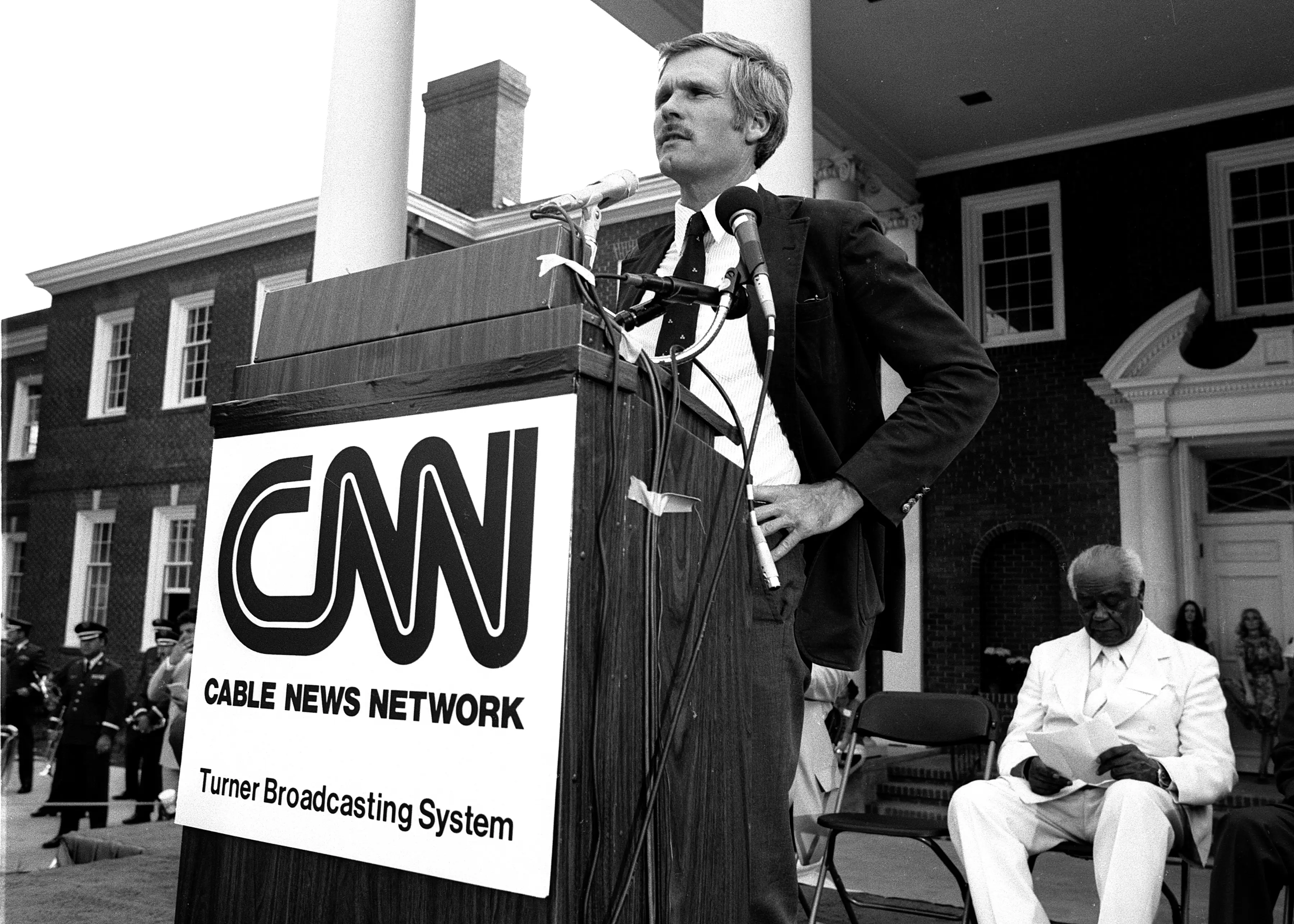 Ted Turner, standing in front of a lectern with a large CNN sign, is putting his hands on his hips and looking into a crowd.