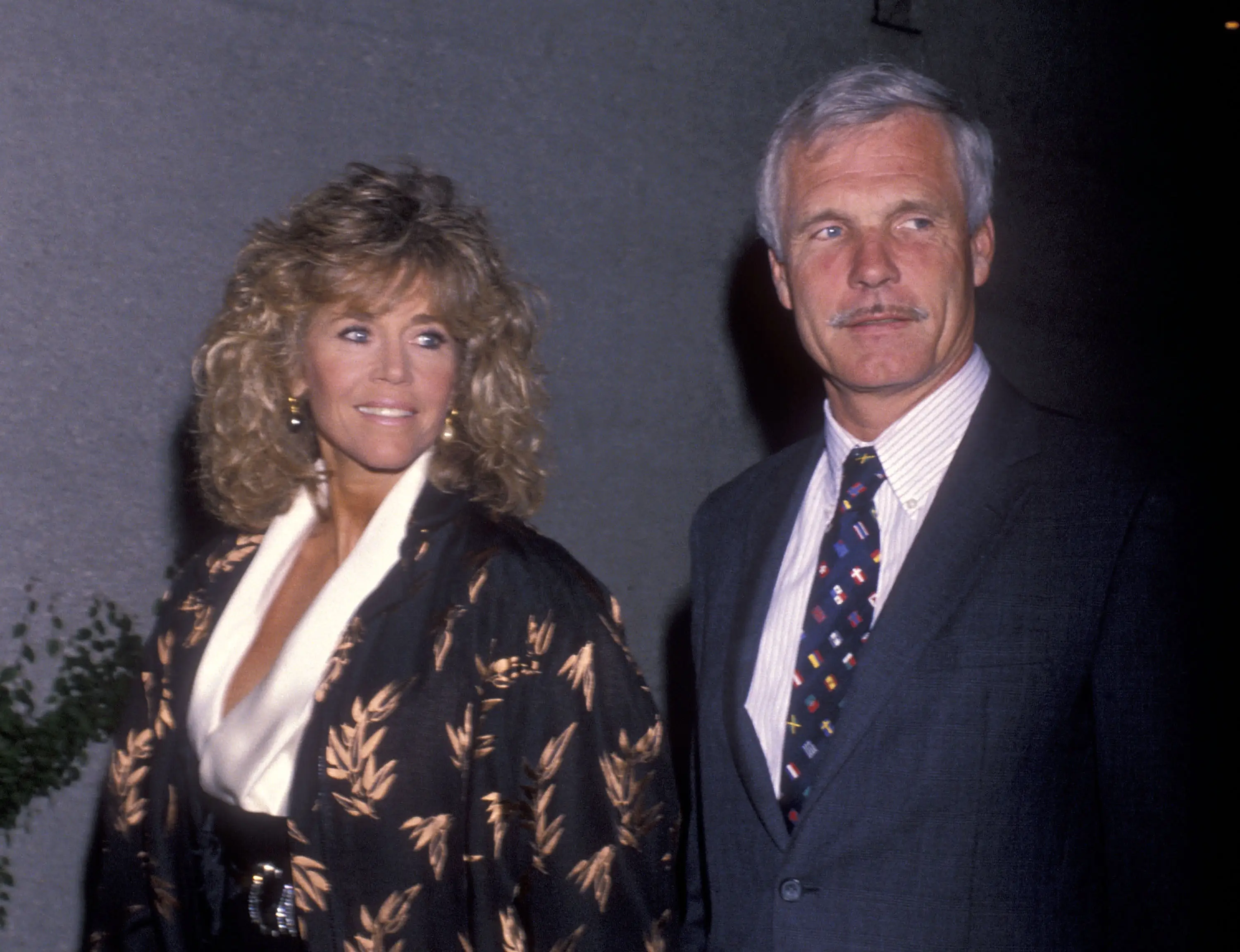 Ted Turner, dressed in a suit, turns toward a camera next to his then-partner, actor Jane Fonda.