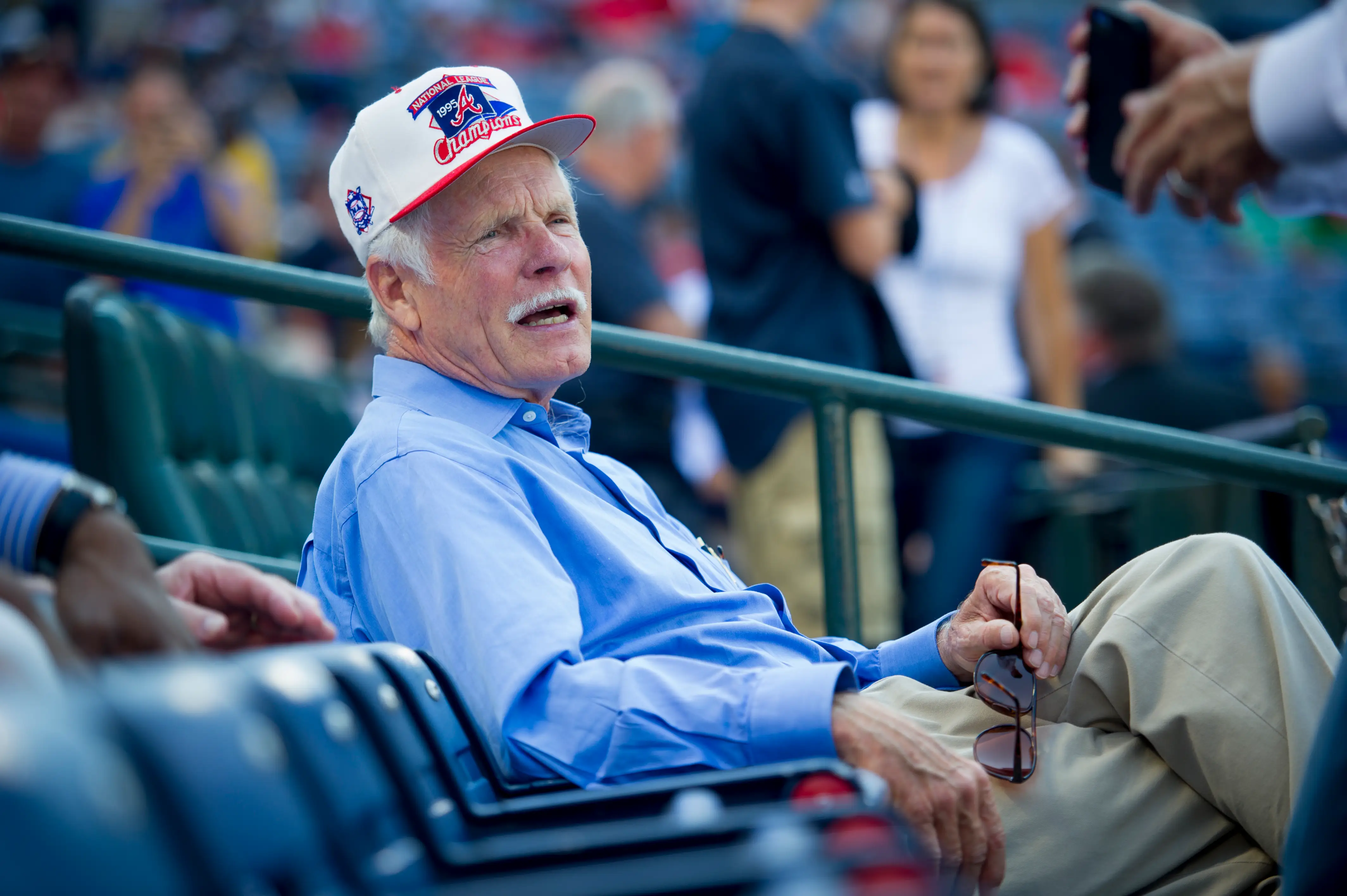 Ted Turner, dressed in a blue button-up shirt and a white flat-brim hat, attends a Major League Baseball game.
