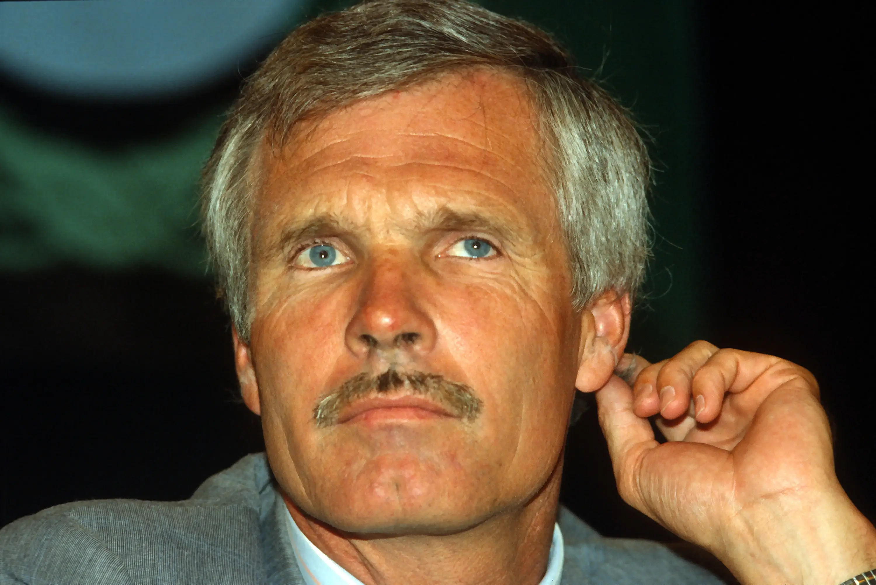 Ted Turner lifts his left hand behind his ear during a press conference.