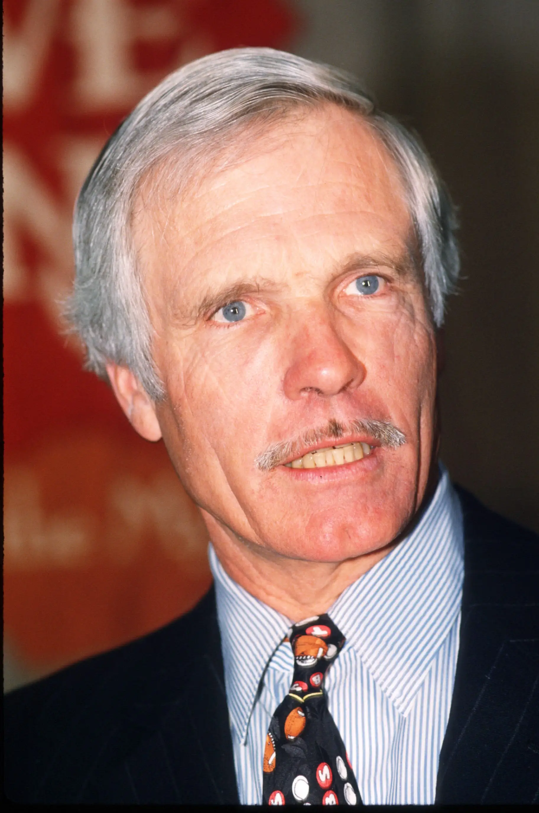 Ted Turner turns to a camera during a press conference. He's wearing a sports-themed tie.