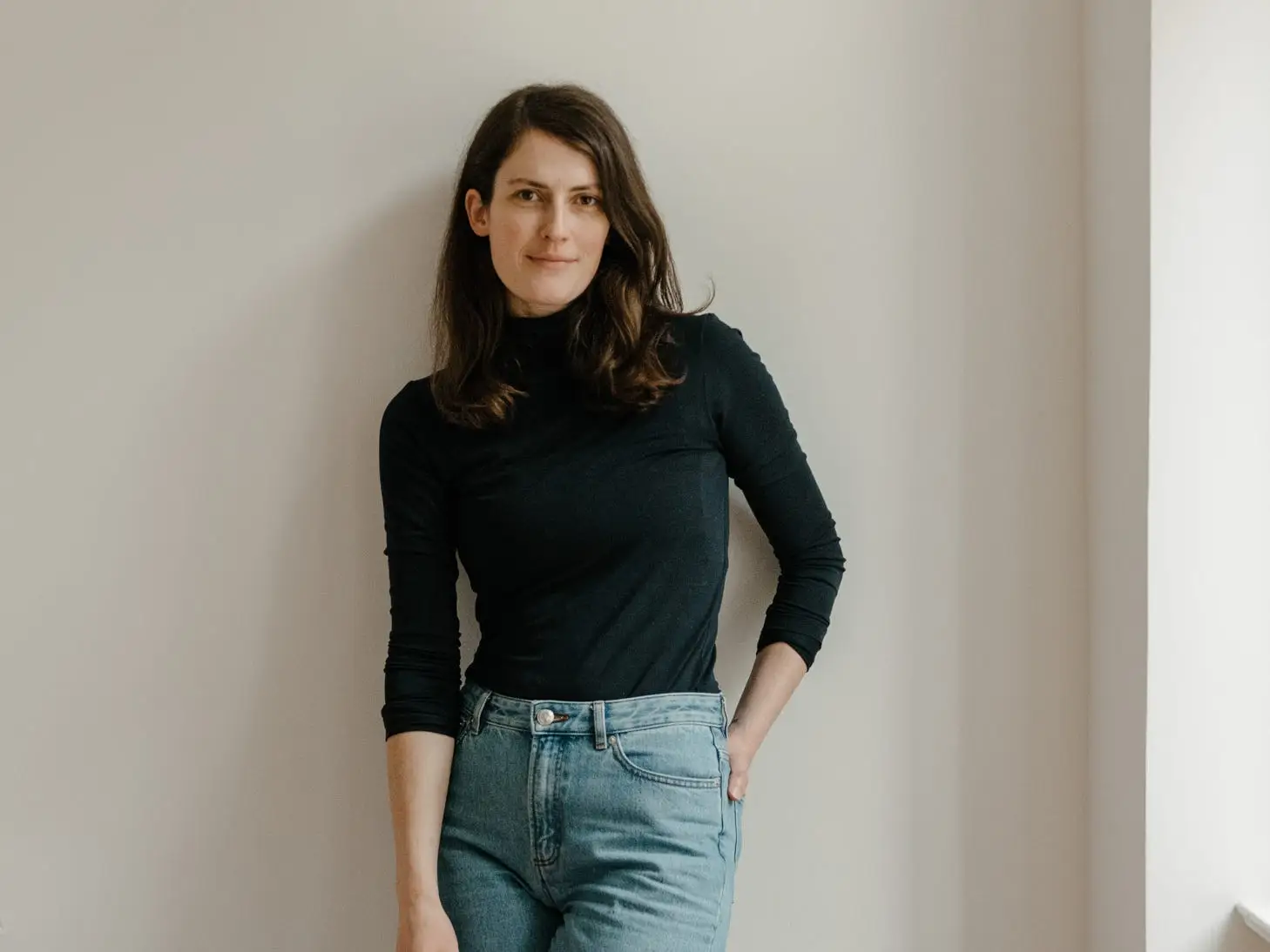 A woman in a black long-sleeved top and jeans leaning against a wall.