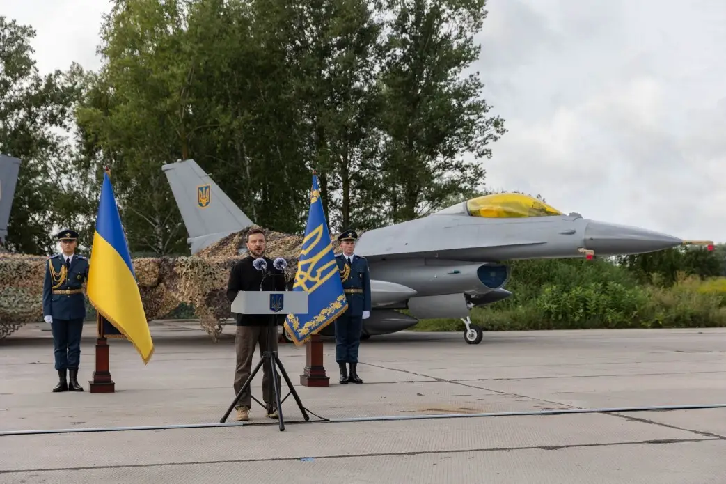 A man in black stands behind a podium with a grey fighter jet on tarmac, two men in military gear, and blue and yellow flags behind him