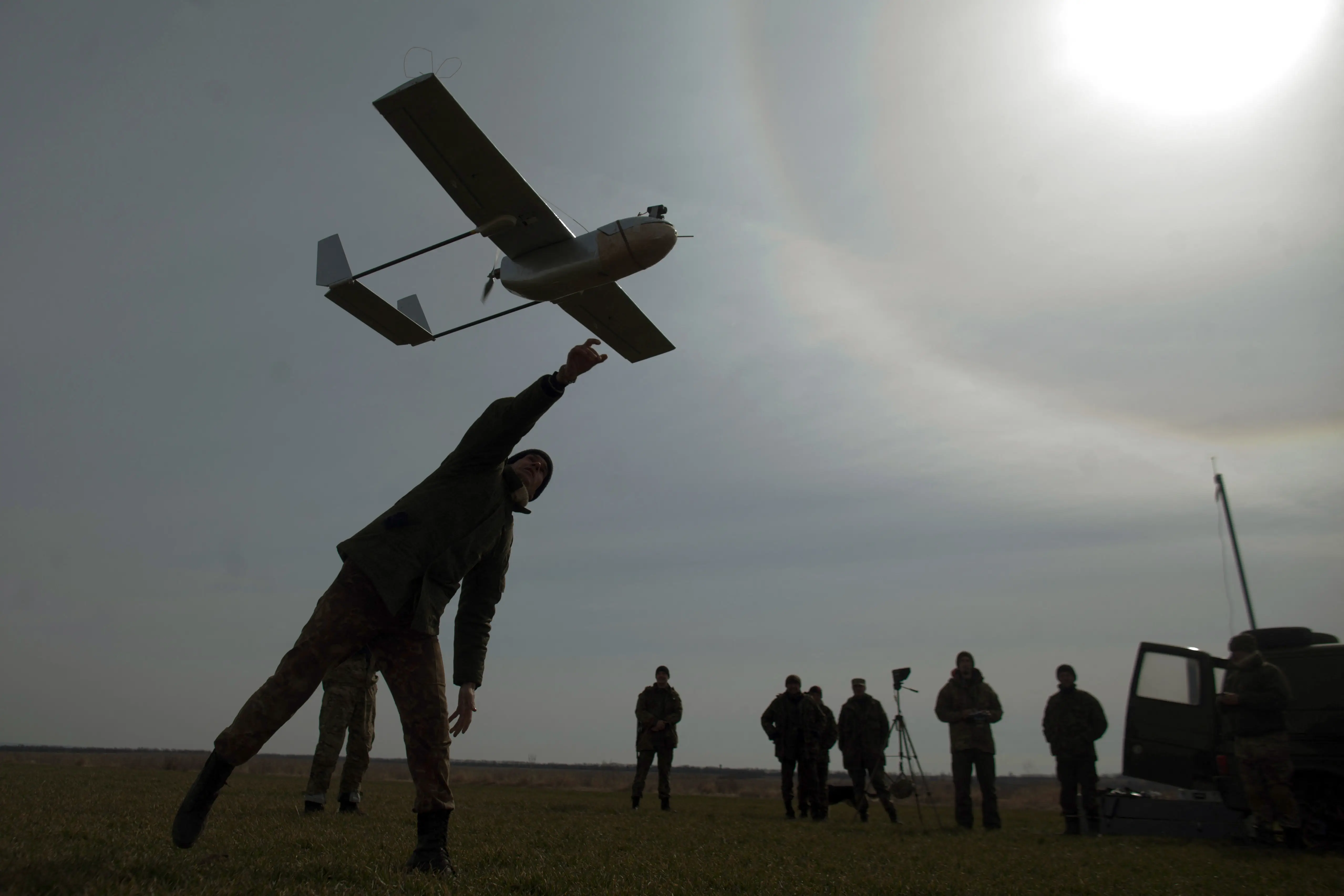A man, silhouetted in the sun, stands and launches a drone in the air with other figures watching on