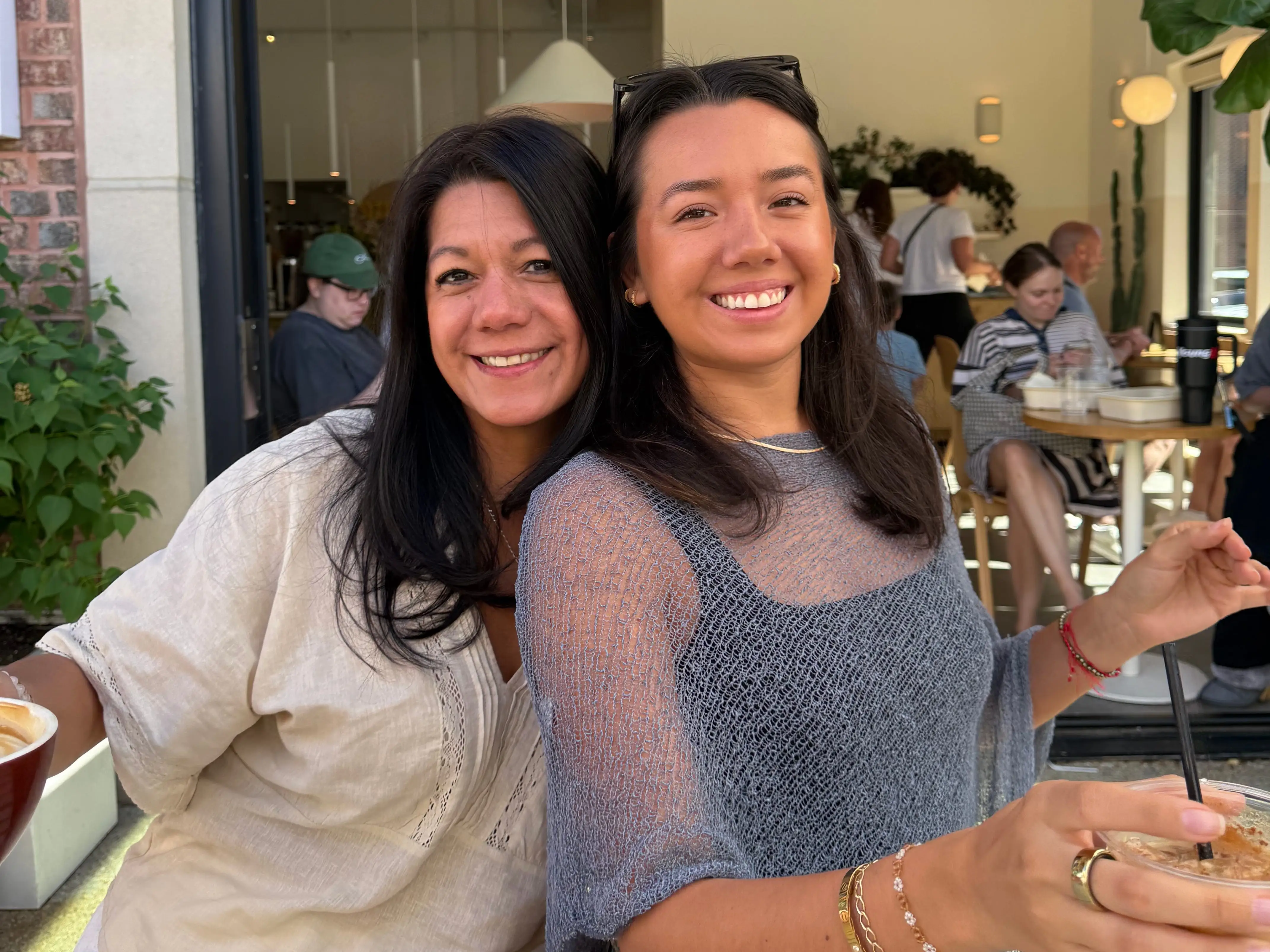 A mom and a daughter smile for a picture while sitting at a restaurant, the daughter has a coffee in her hand.