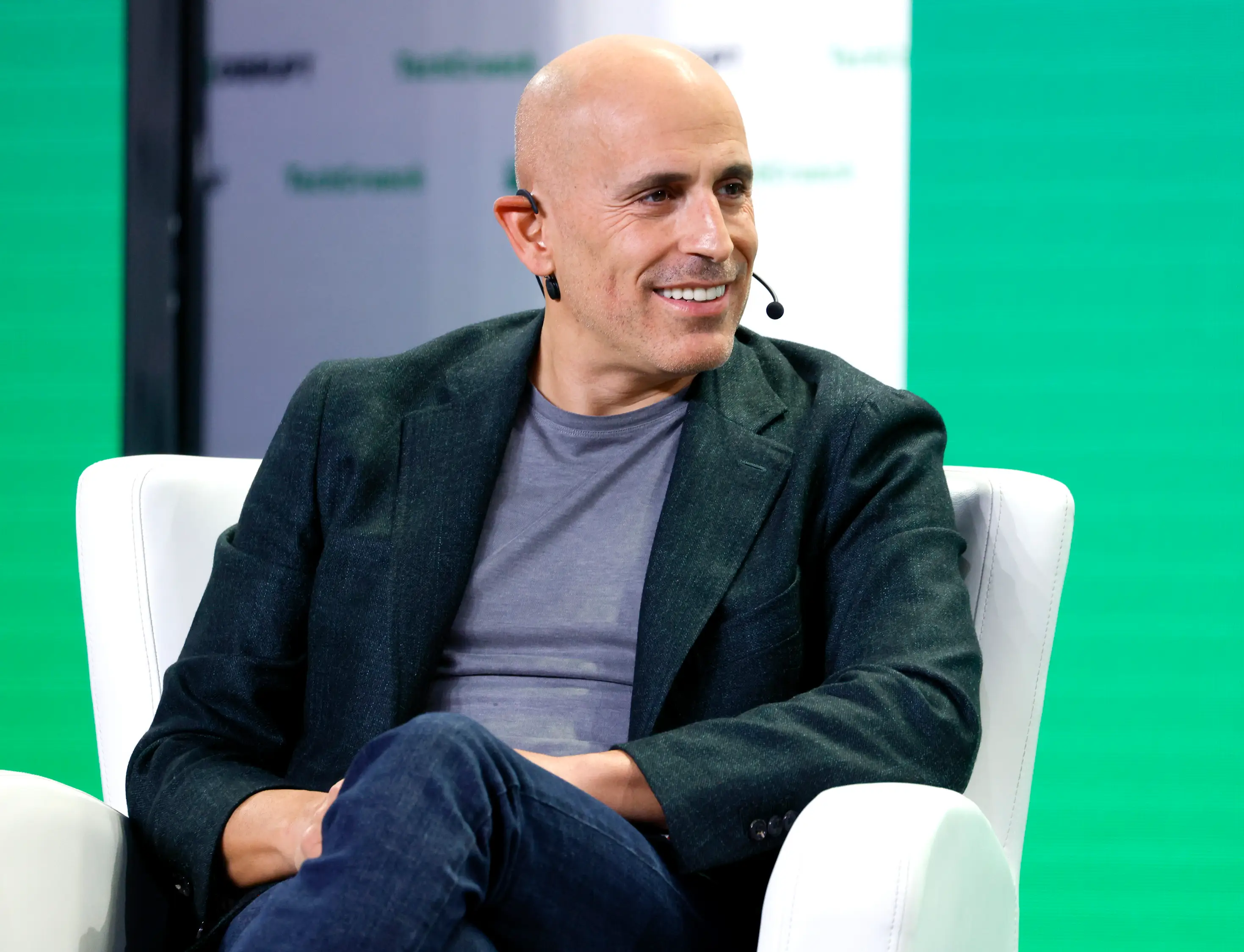 Marc Lore sits in a white chair on a conference stage wearing a dark grey blazer, grey t-shirt, blue jeans and a microphone while listening to a presenter.