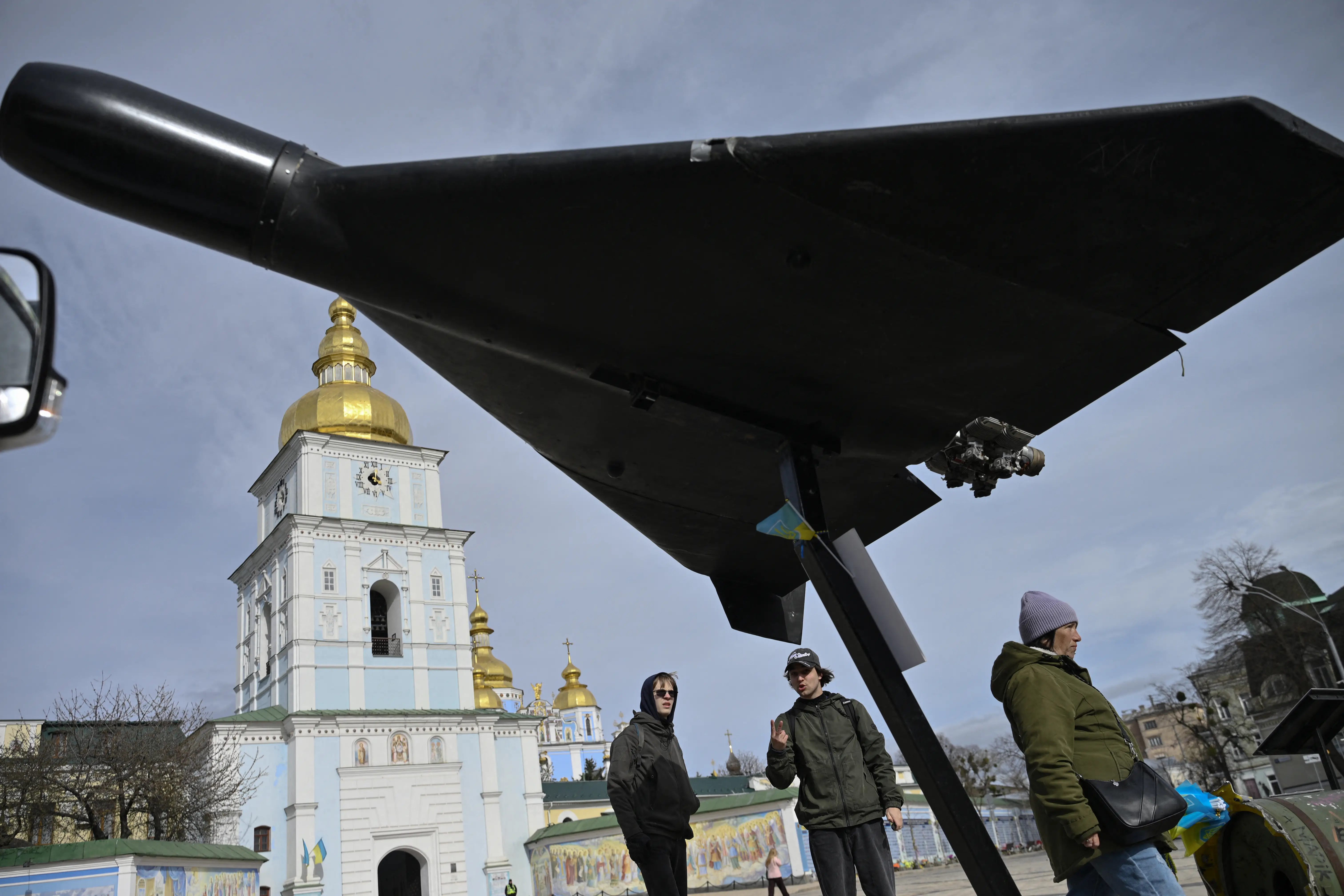 People look up at a large black drone on display in front of a blue and white church with a gold top