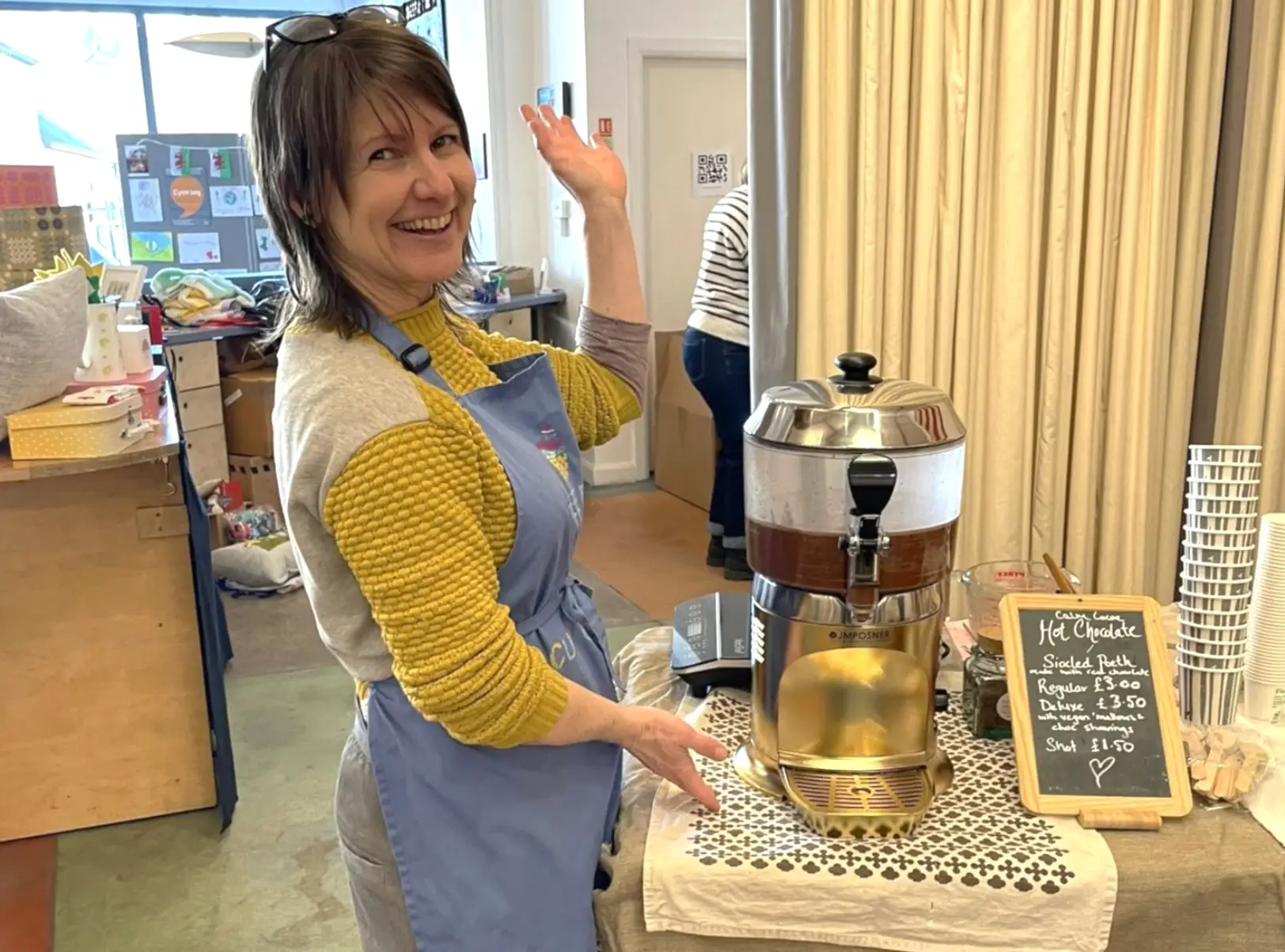 The writer posing and waving in front of a hot chocolate machine.