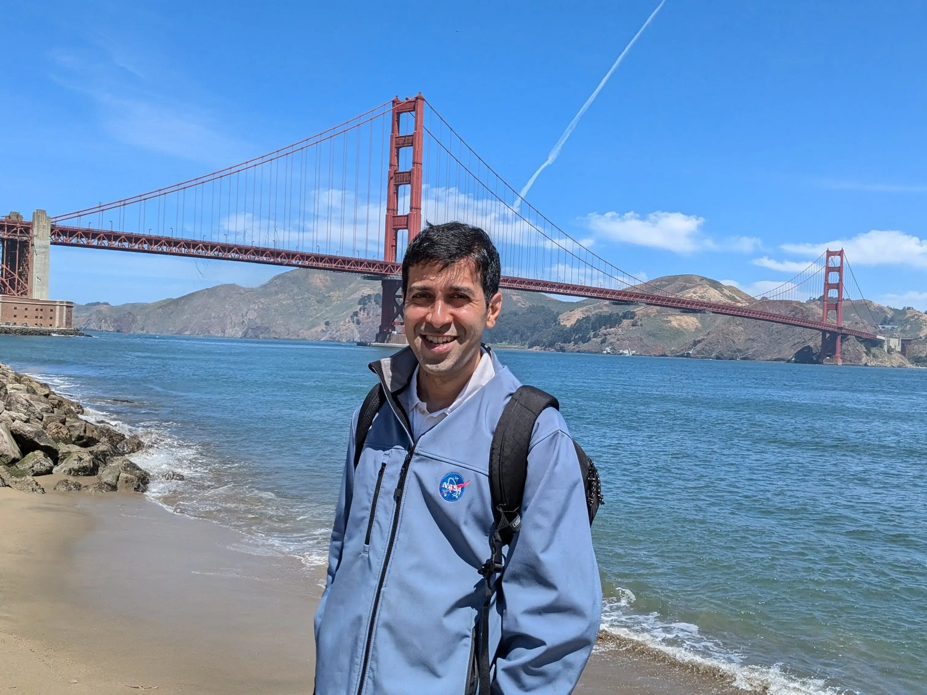 Anand Ramanathan is standing in front of the ocean, wearing a blue jacket with the NASA logo on it.
