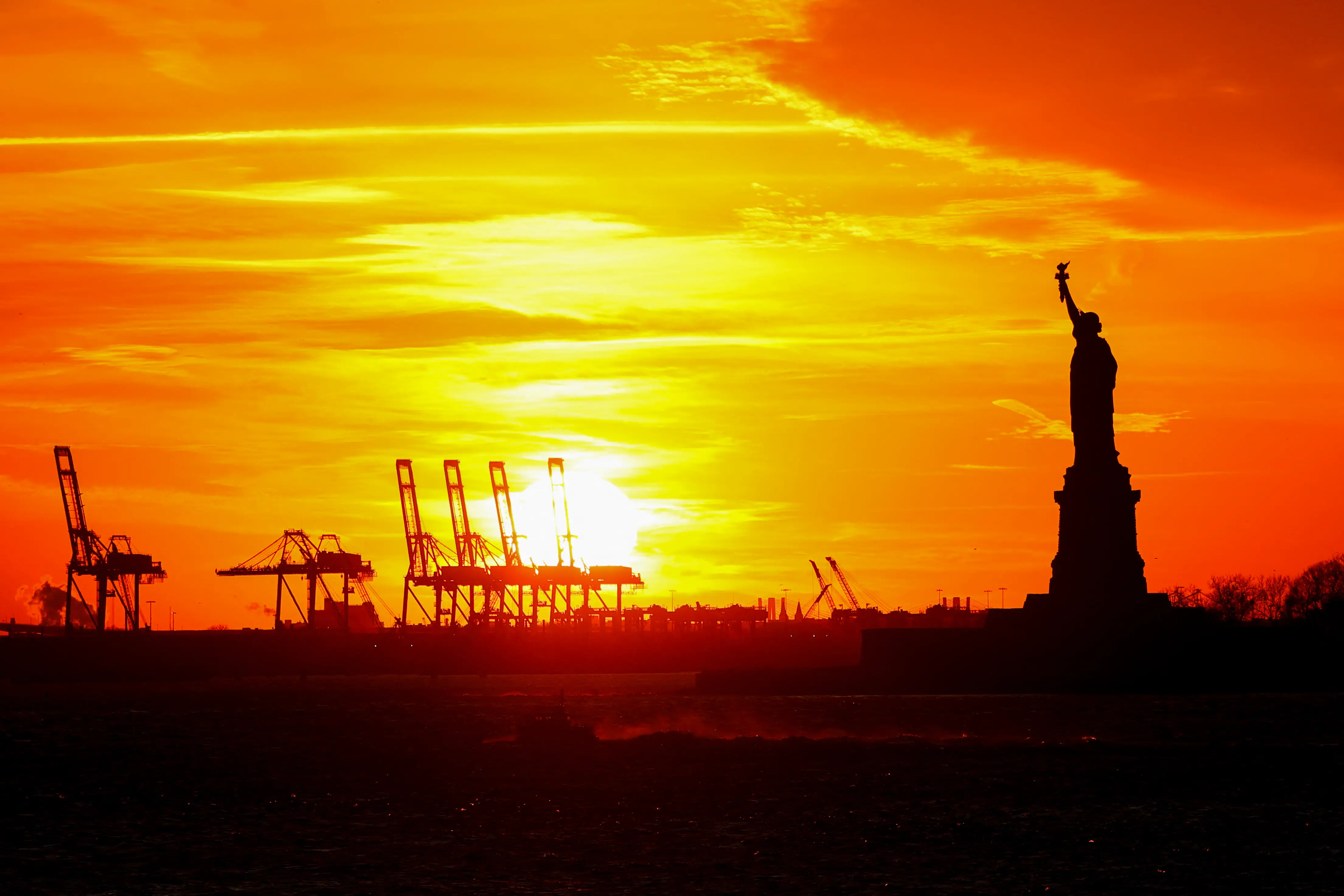 Cranes for handling shipping containers stand in the far distance as the Statue of Liberty stands in the middle distance as the sun sets near New York City.