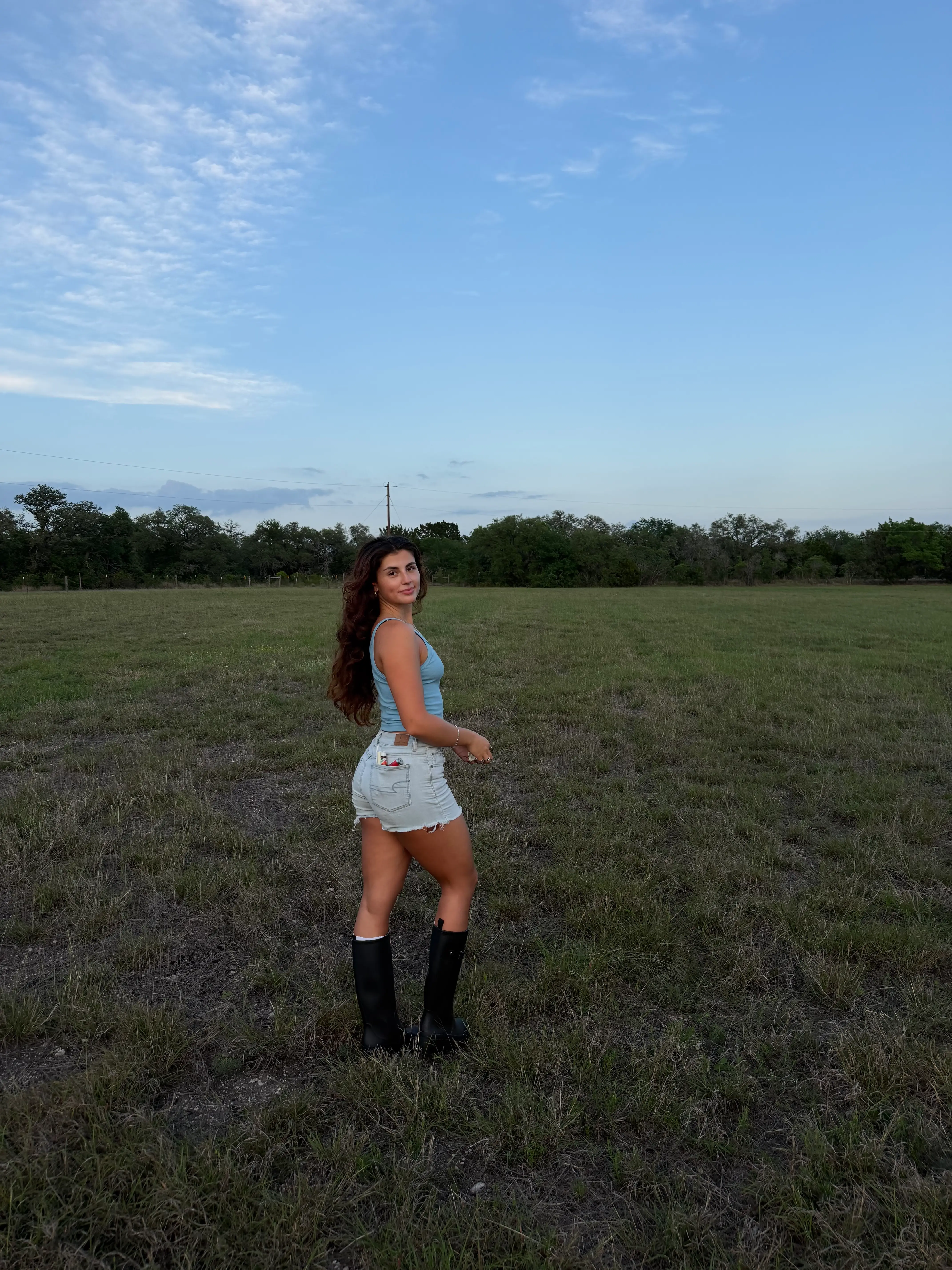A woman wearing a tank top, shorts, and boots stands in a field with a blue sky behind her.