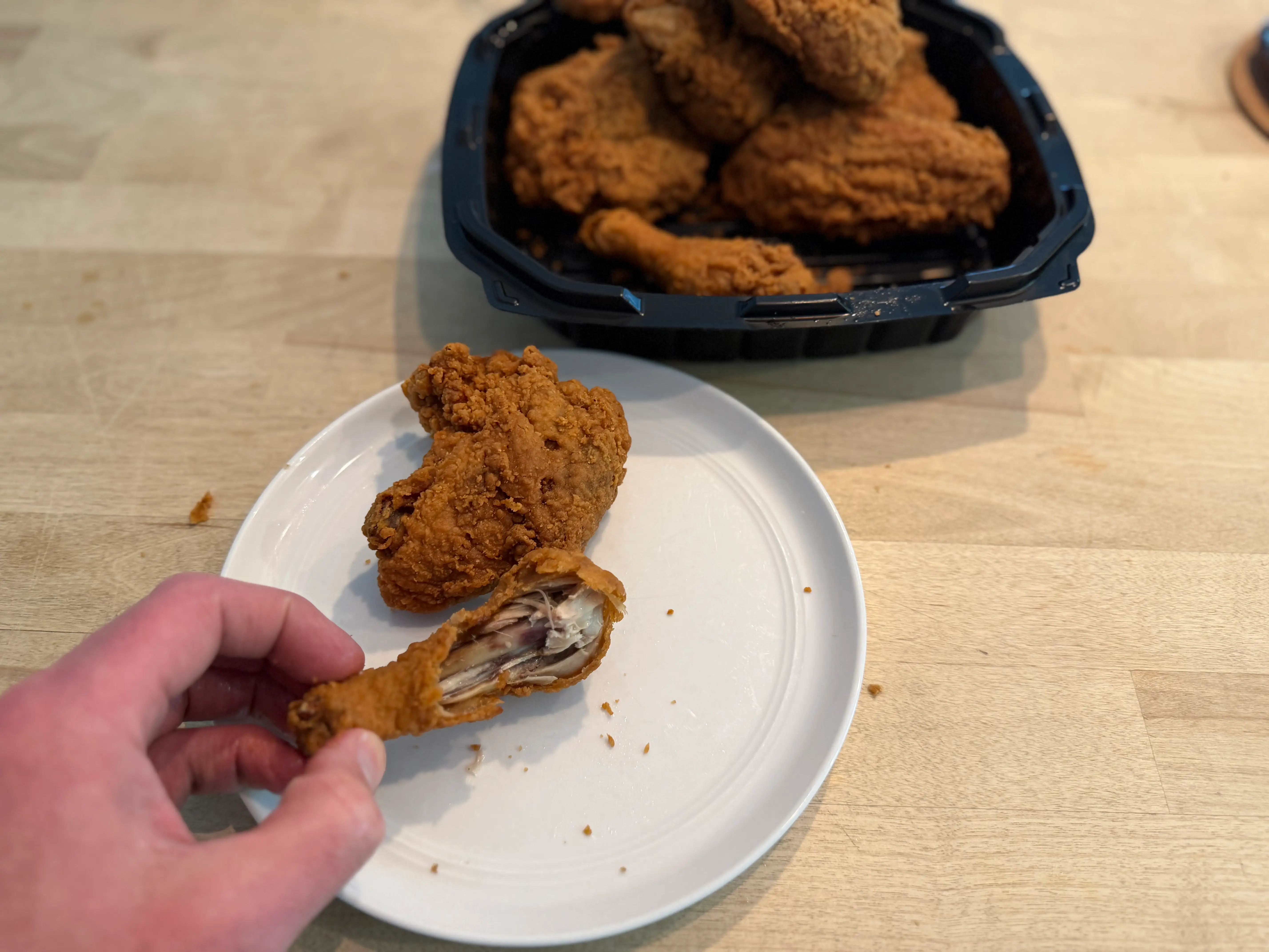Hand holding piece of Walmart fried chicken on plate with container of more chicken behind it