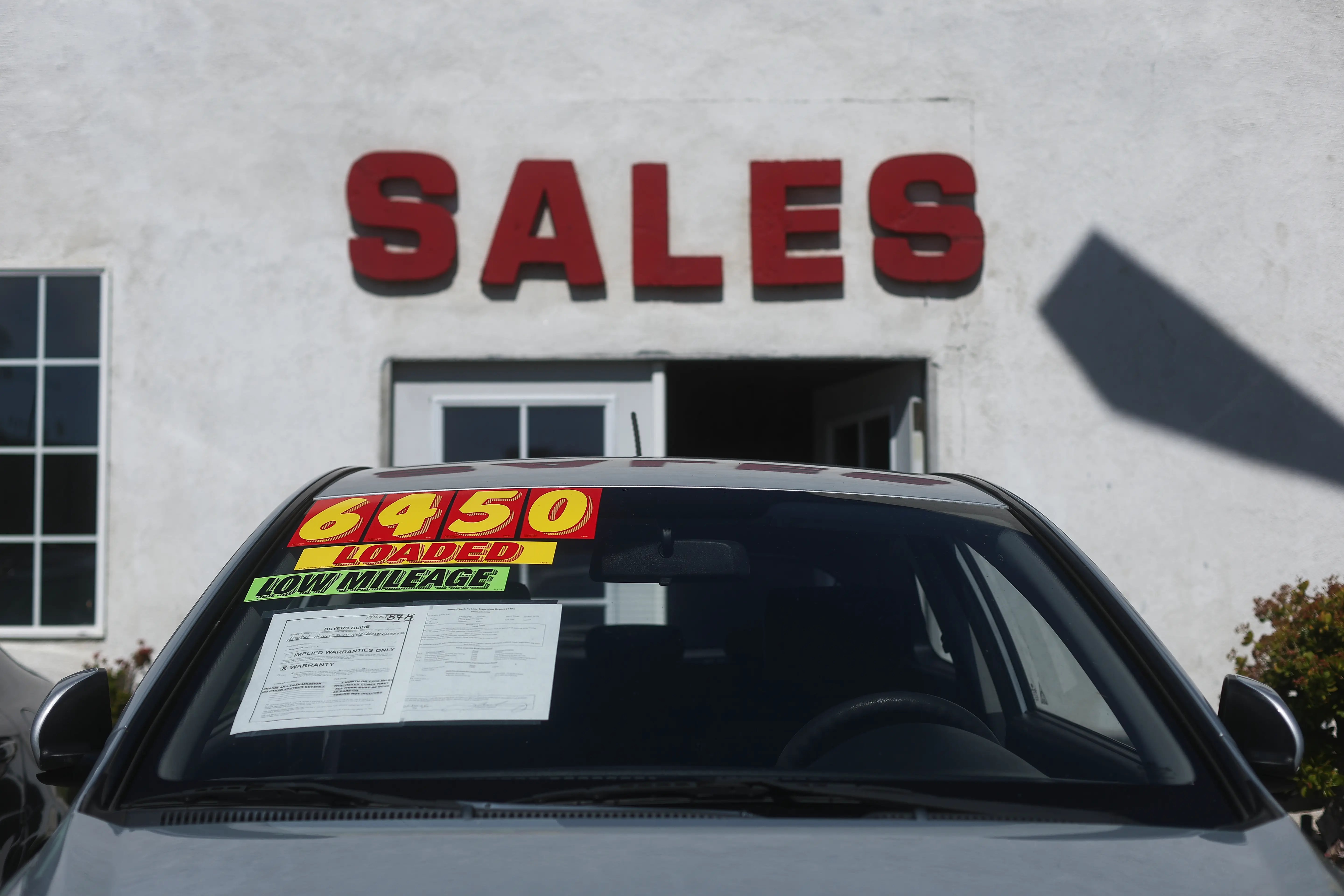 A used car with a $6,450 sticker price is parked on a dealership lot in front of the sales entrance door.