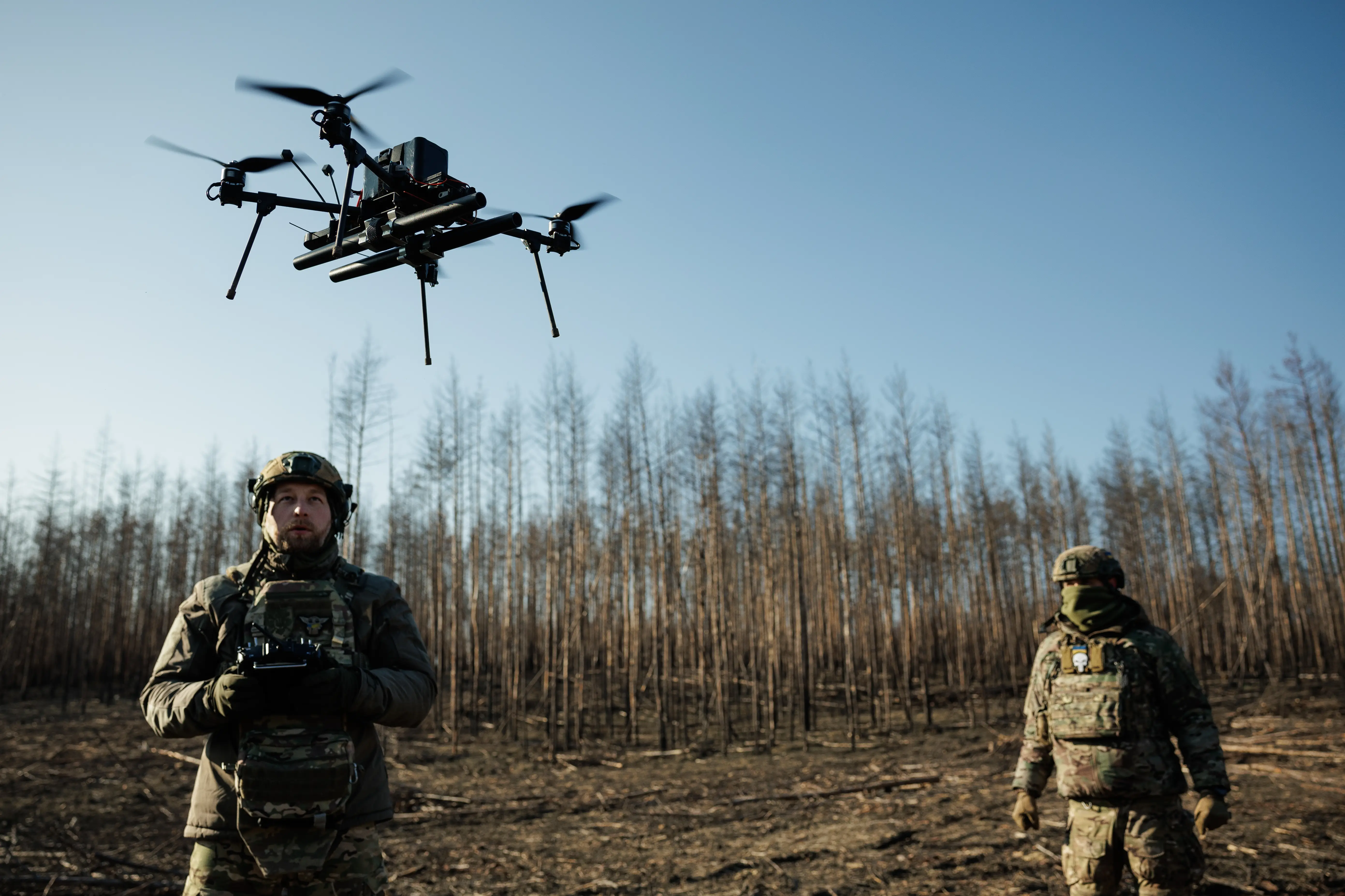 Two figures in camouflage gear stand in a field with bare trees and a blue sky behind them with a drone hovering above