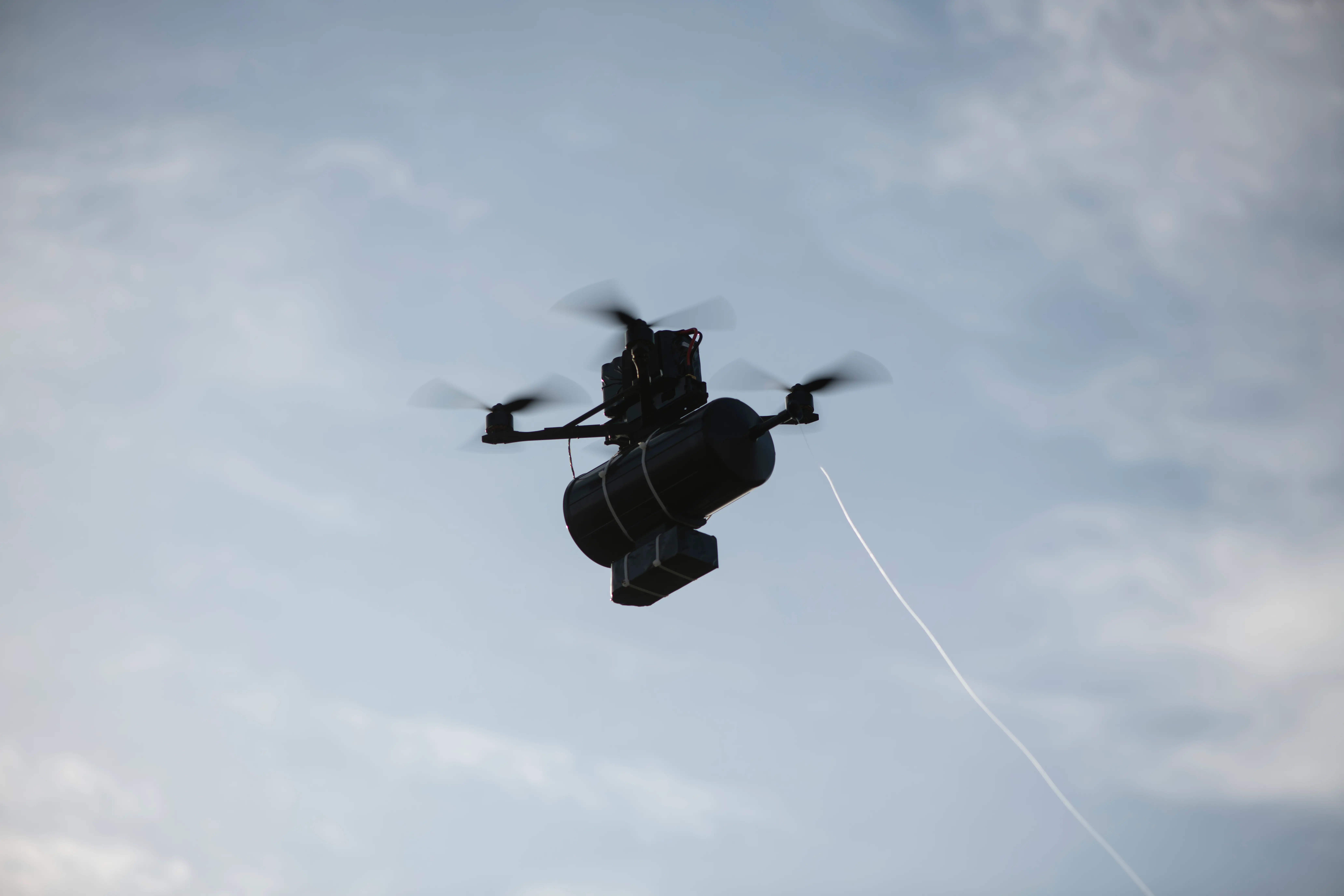 A small black drone in a blue and cloudy sky with a thin white wire coming down from inside it