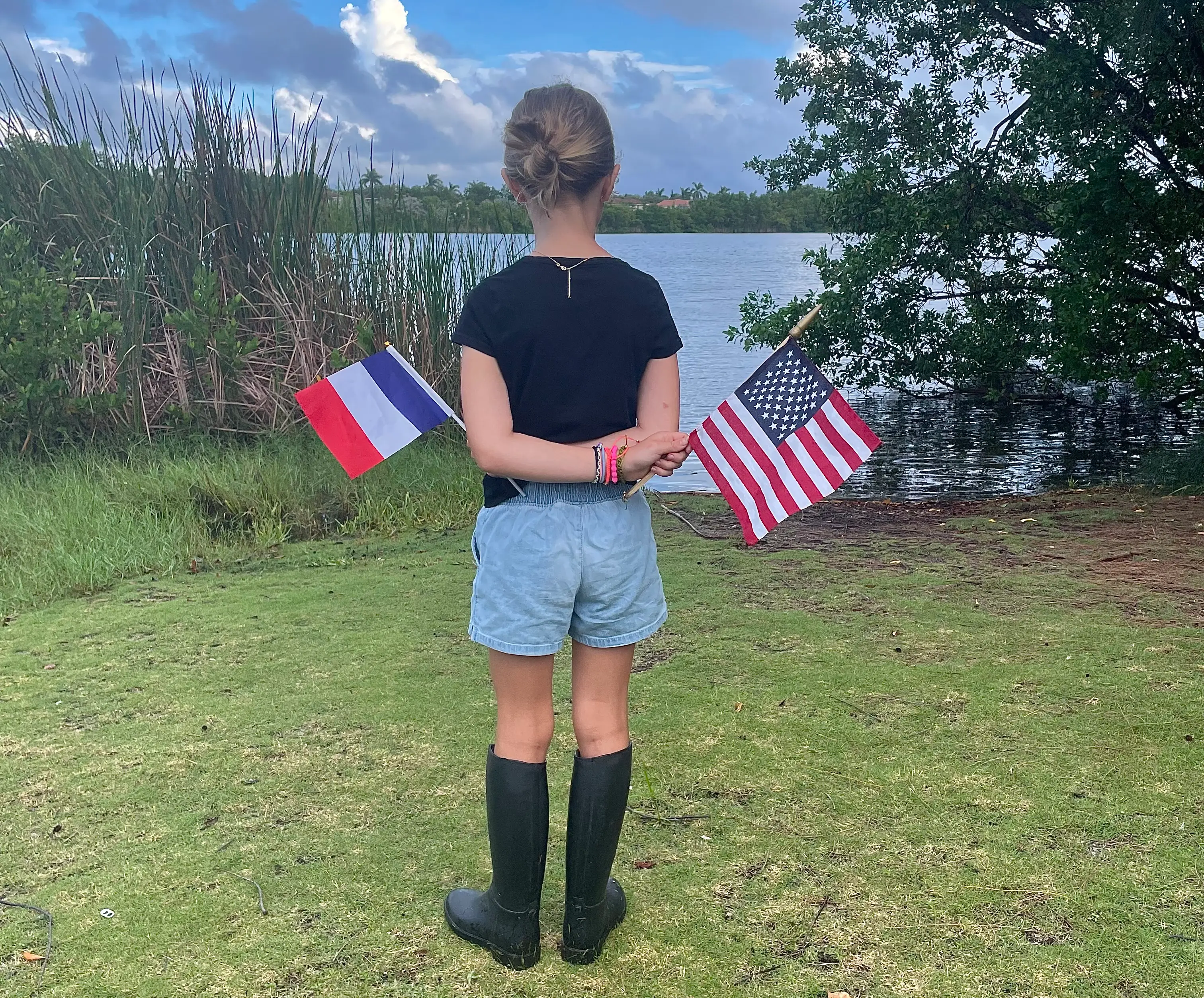 The author's daughter standing near a body of water, holding an American flag in one hand and a French flag in another.